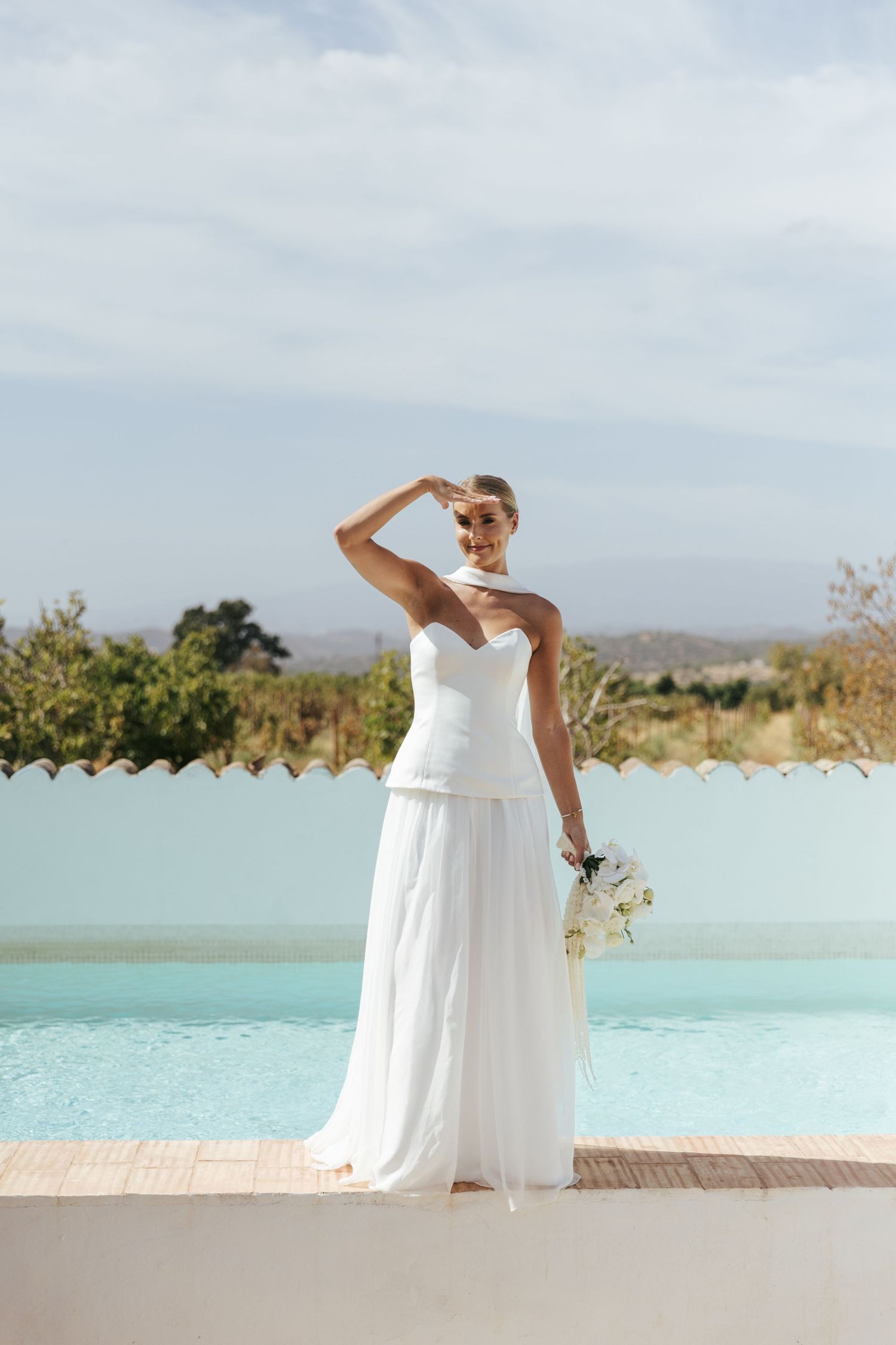 Bride in white strapless gown holding bouquet by pool, shielding eyes from sun with scenic landscape backdrop.