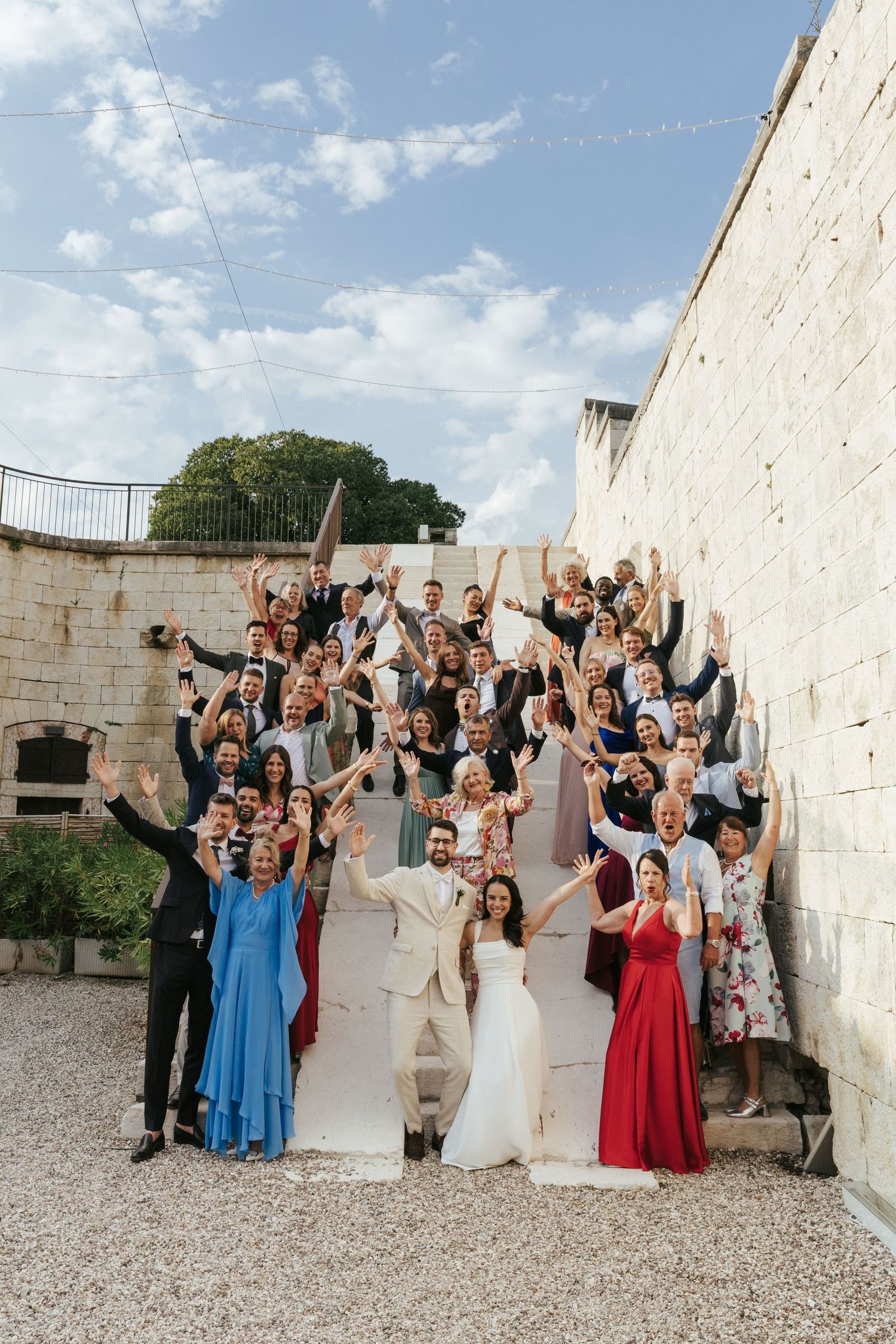 Large group of wedding guests pose for a celebratory photo against white stone walls outdoors.