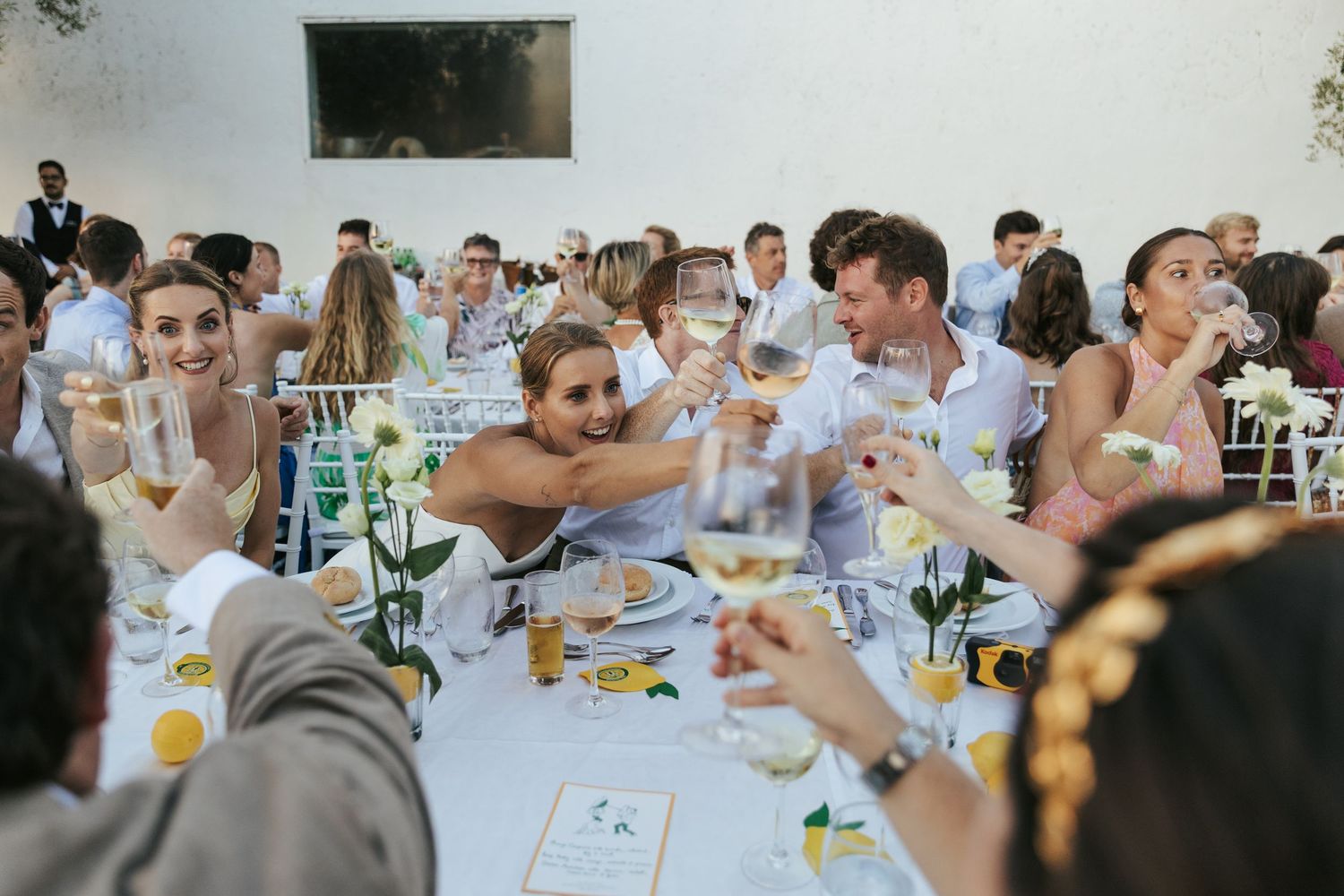 Wedding guests raising glasses in a joyful toast at an elegant outdoor reception with white tablecloths and yellow roses.