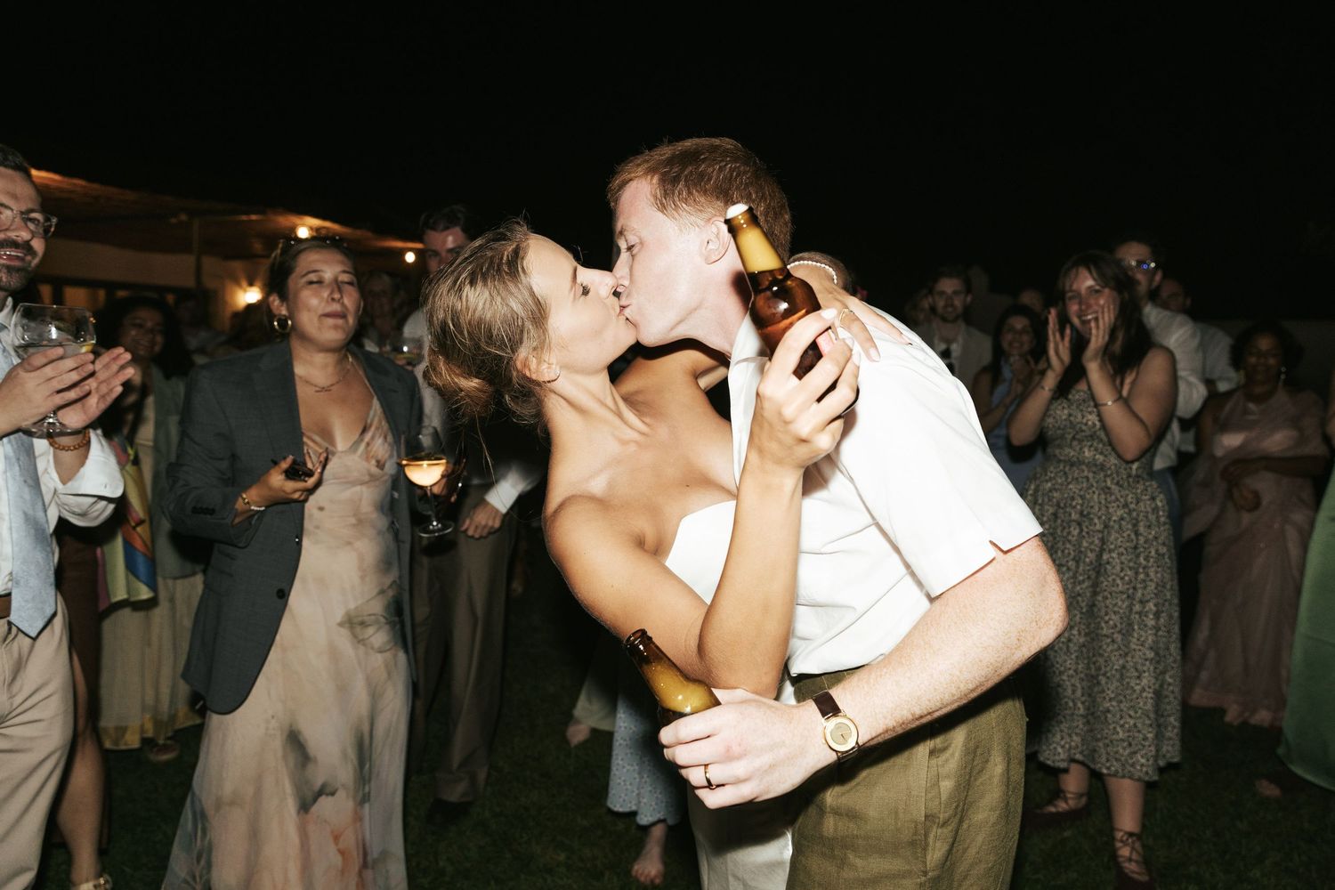 Couple sharing a romantic kiss at outdoor wedding reception, holding beer bottles, guests celebrating in background at night.
