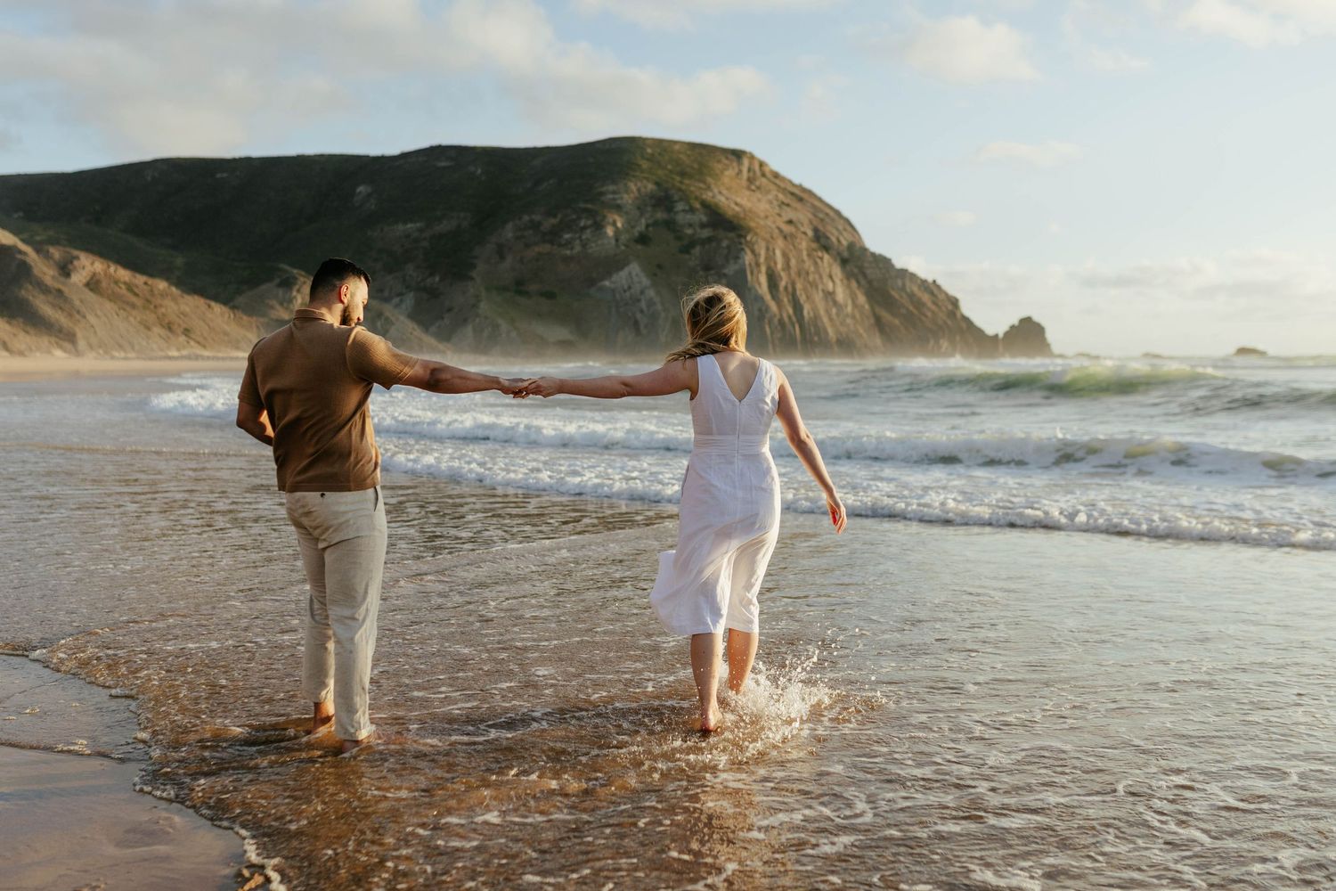 Two people walking hand in hand along a scenic beach shoreline with dramatic coastal cliffs in the background at sunset.