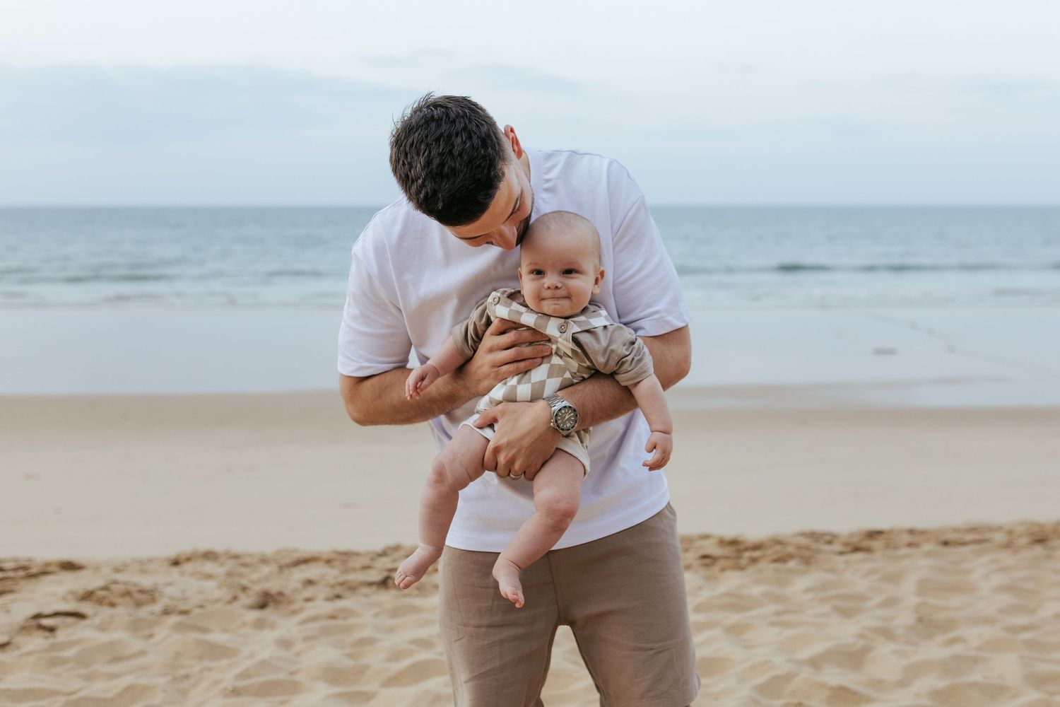 A tender moment shared between parent and baby on a sandy beach with ocean waves in the background.