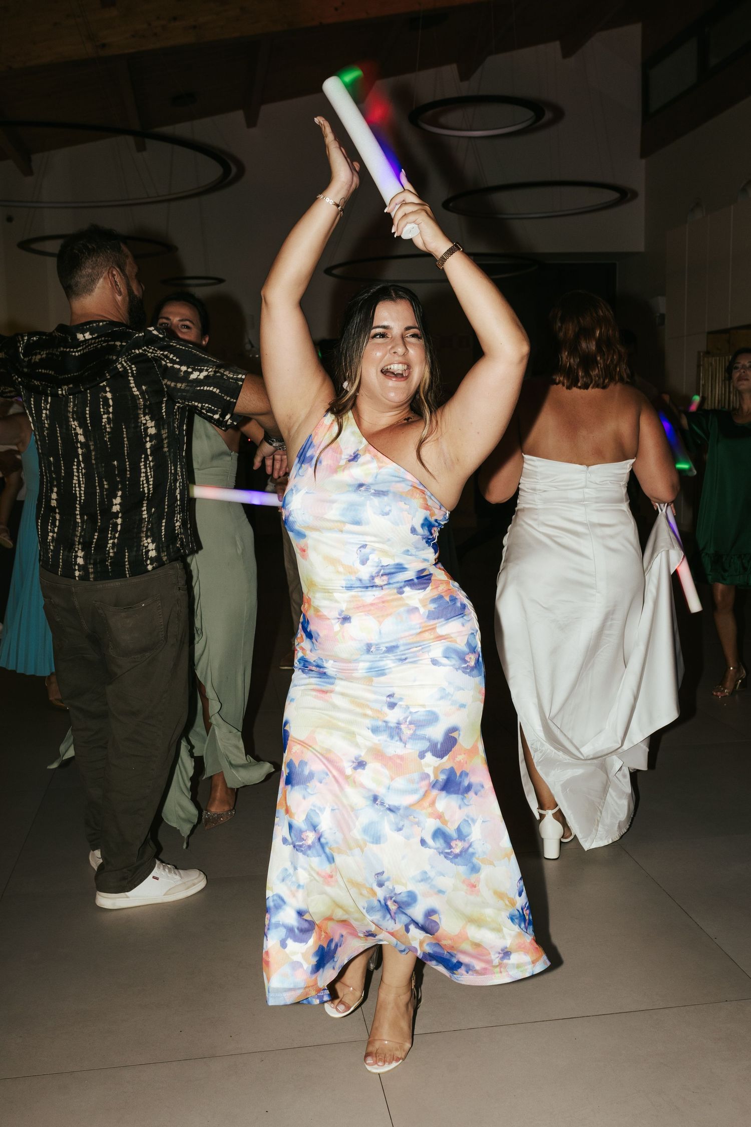 A guest in a floral dress dances joyfully on the reception floor while holding light-up party sticks.