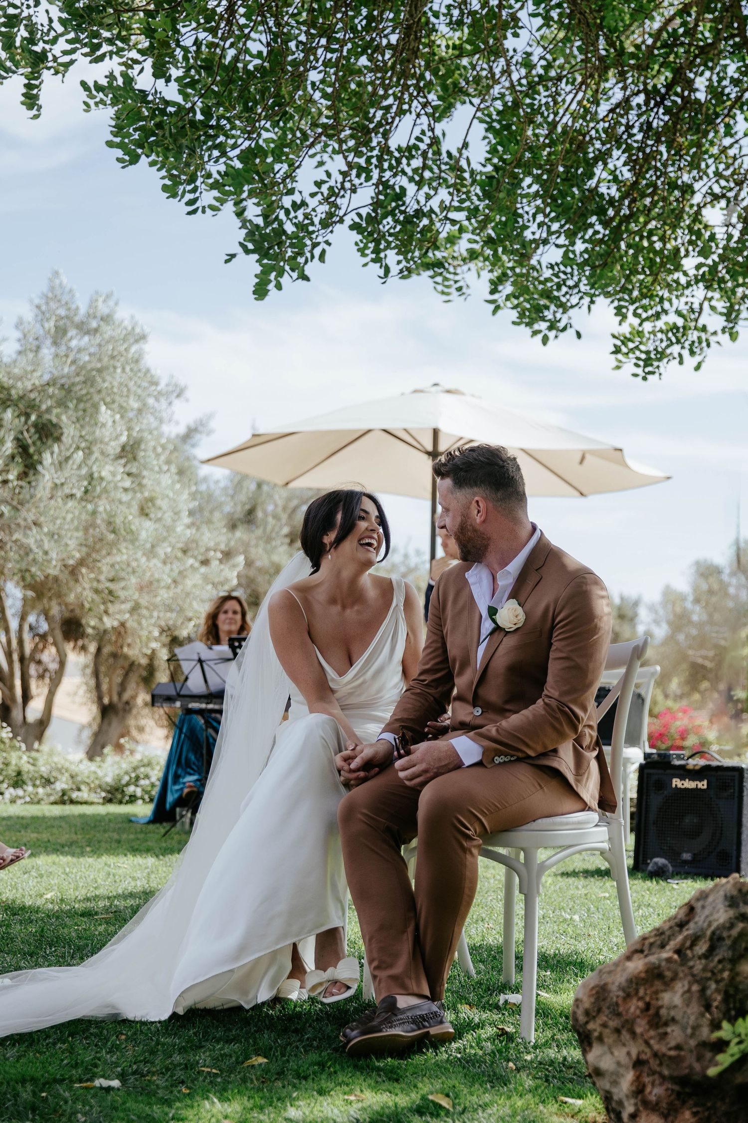 A couple shares an intimate moment seated beneath a white umbrella in a sun-dappled garden setting.