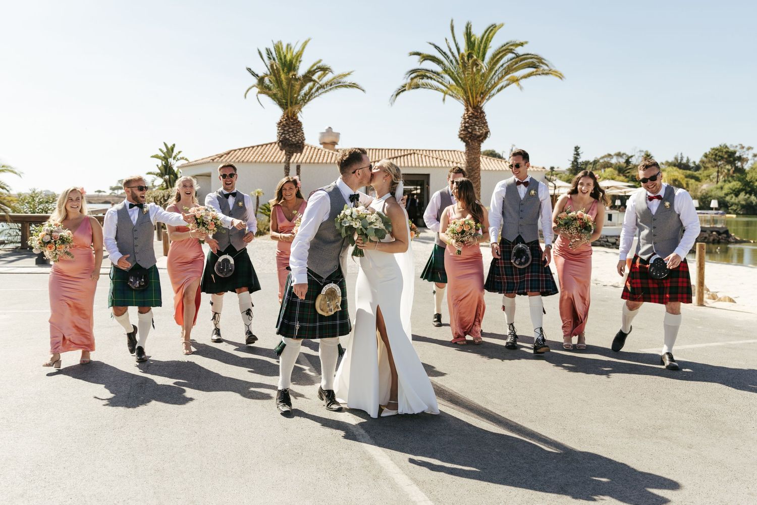 Wedding party members walk in a line along a sunny outdoor path with palm trees in the background.