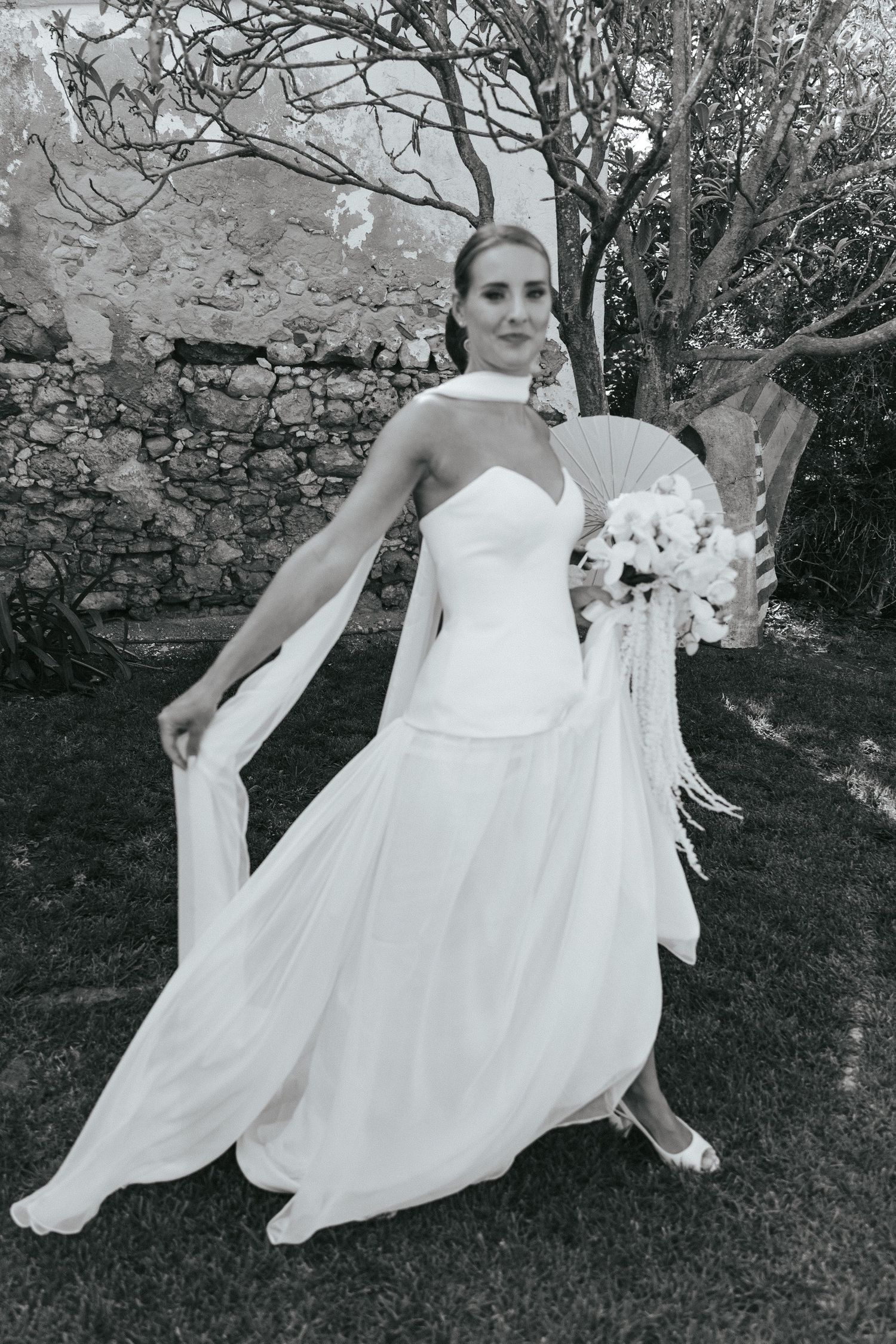 Black and white photo of a bride in a white gown holding a bouquet, posing outdoors near a stone wall.