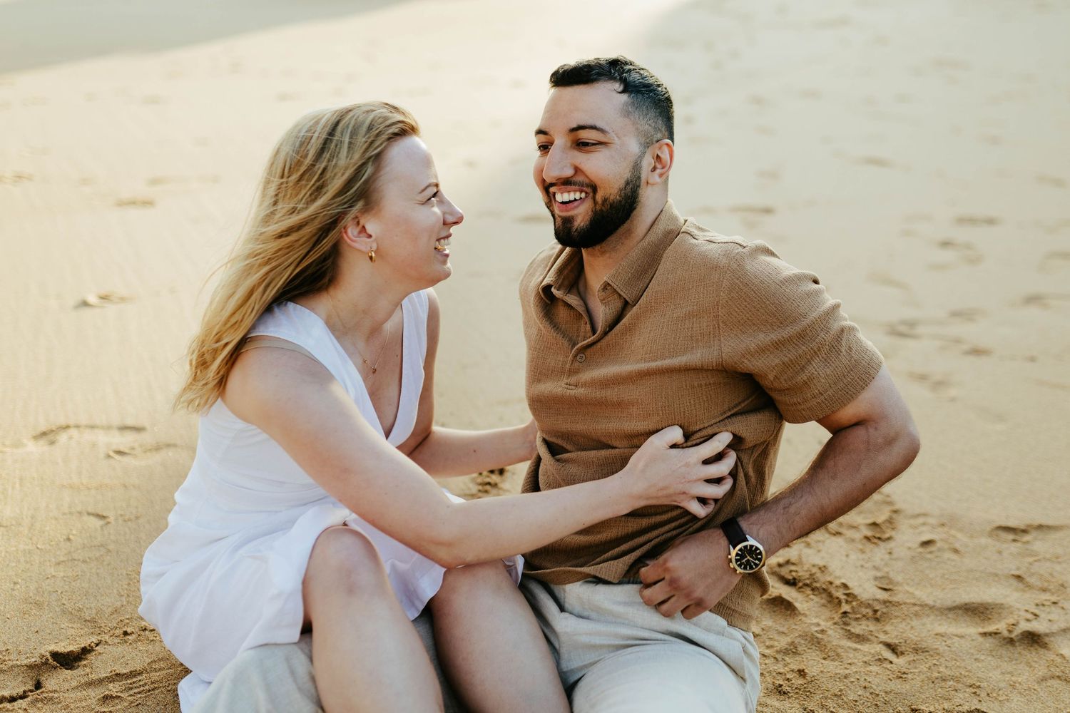 A joyful couple sitting together and laughing on a sandy beach during a casual outdoor photo session.