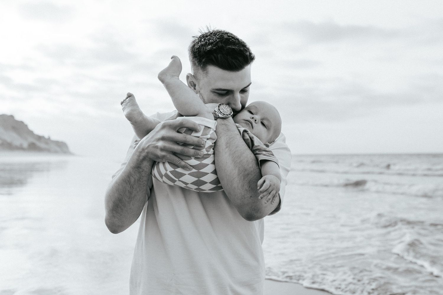 A parent cradles and kisses a baby while standing on a beach in black and white.