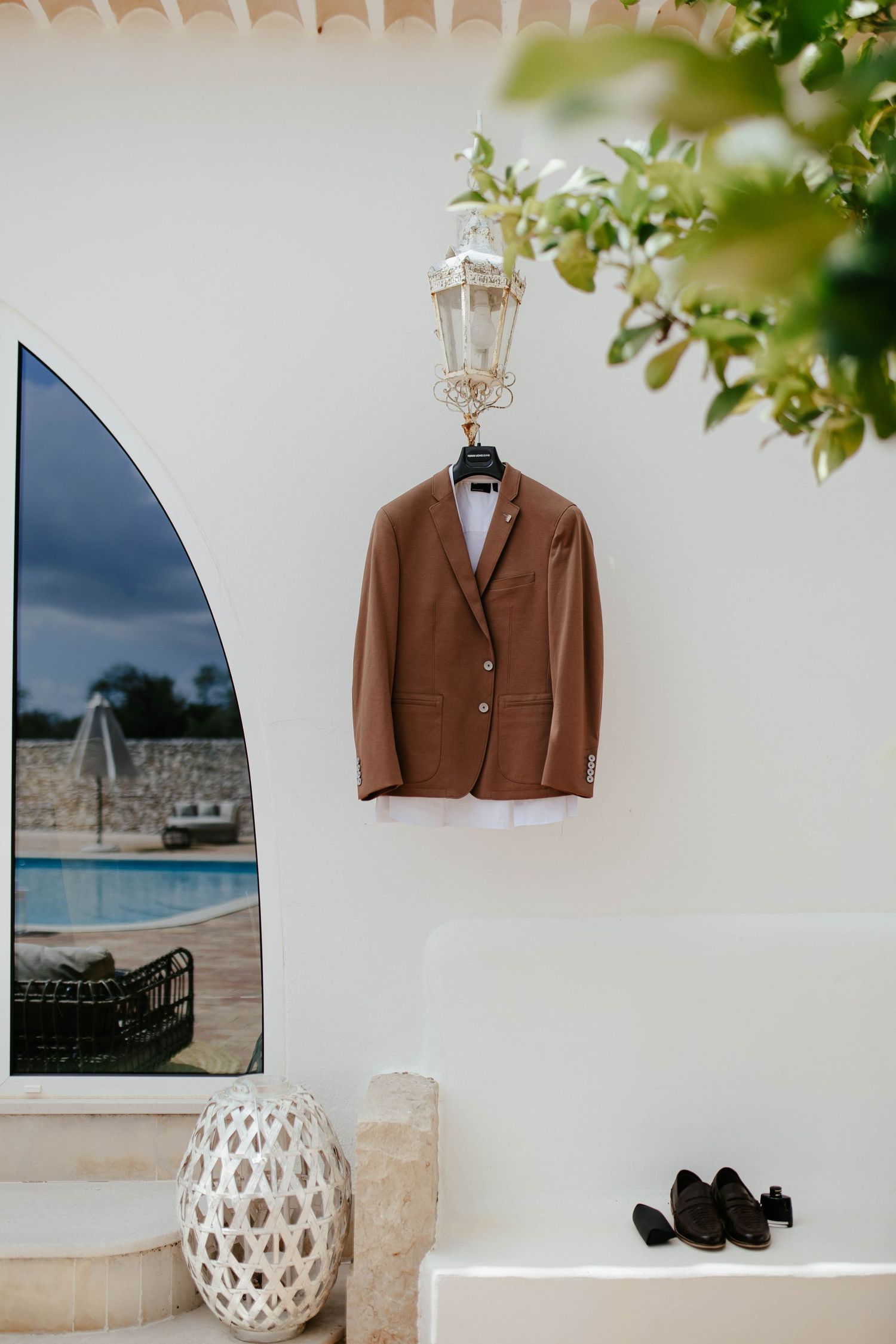 Brown suit jacket hanging on white wall near poolside archway with decorative lantern and woven basket.