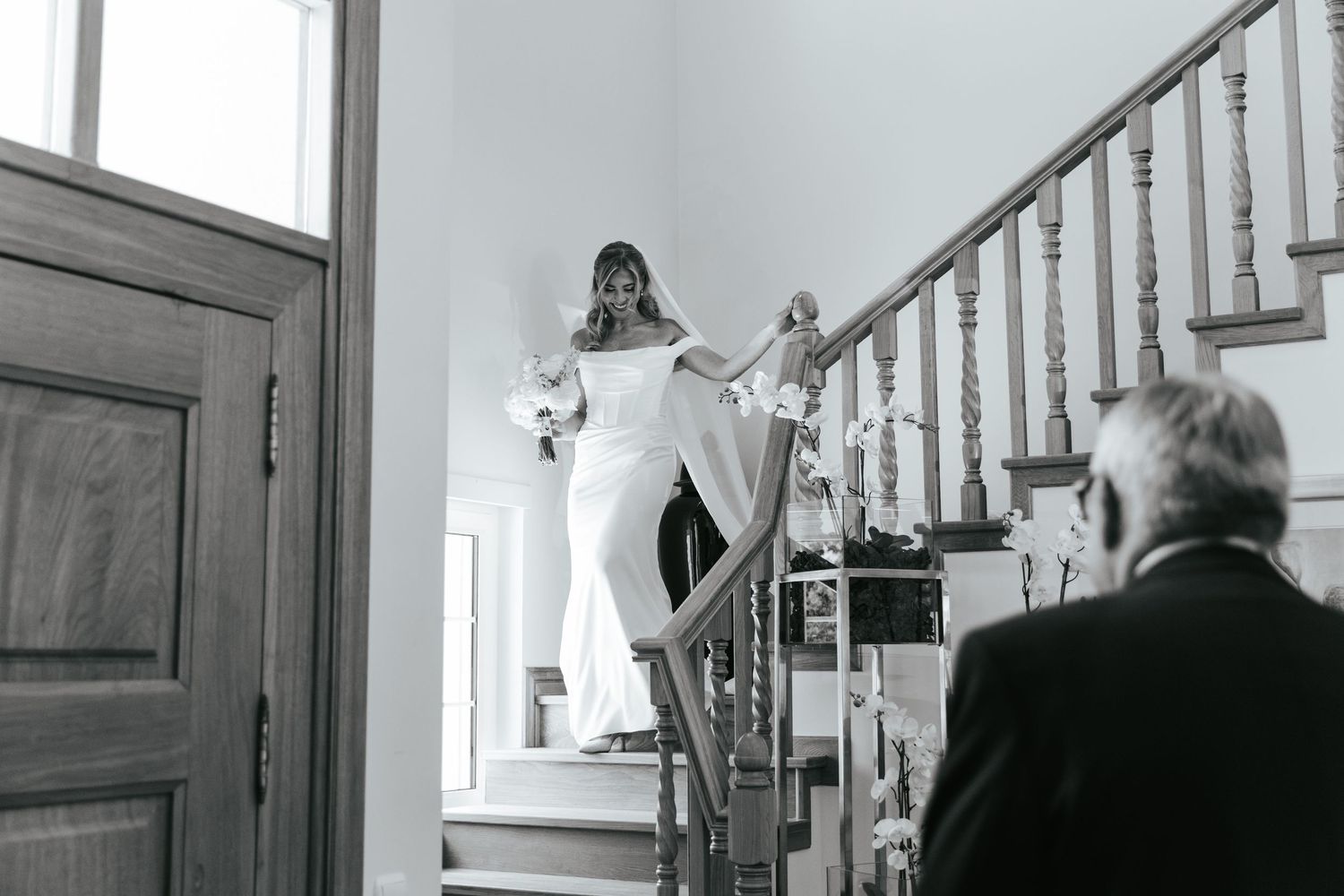 Bride descends staircase in elegant white gown holding bouquet as man watches in black and white wedding photo.