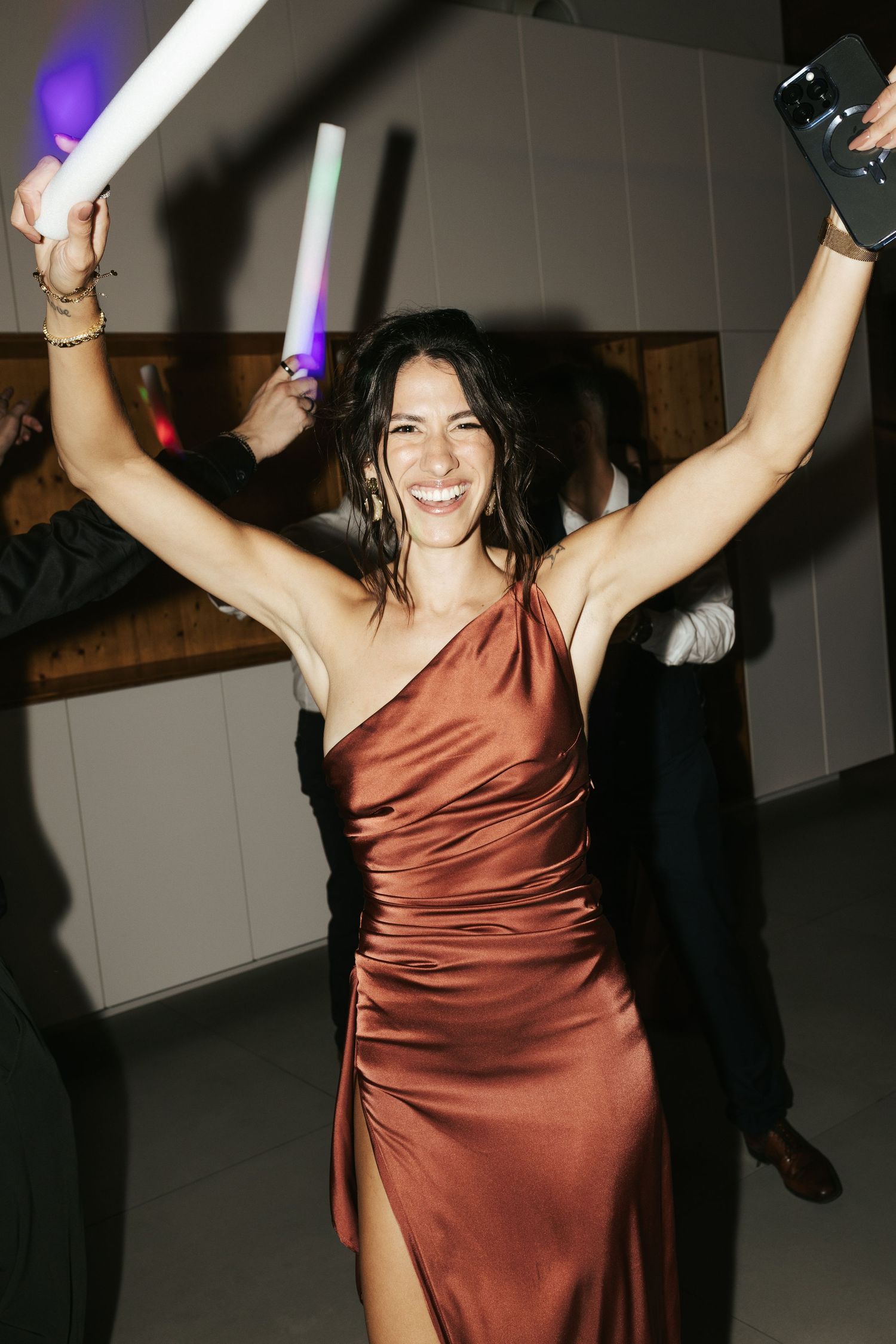 Person wearing copper-colored satin dress with slit dancing joyfully at a party with arms raised under colored lights.
