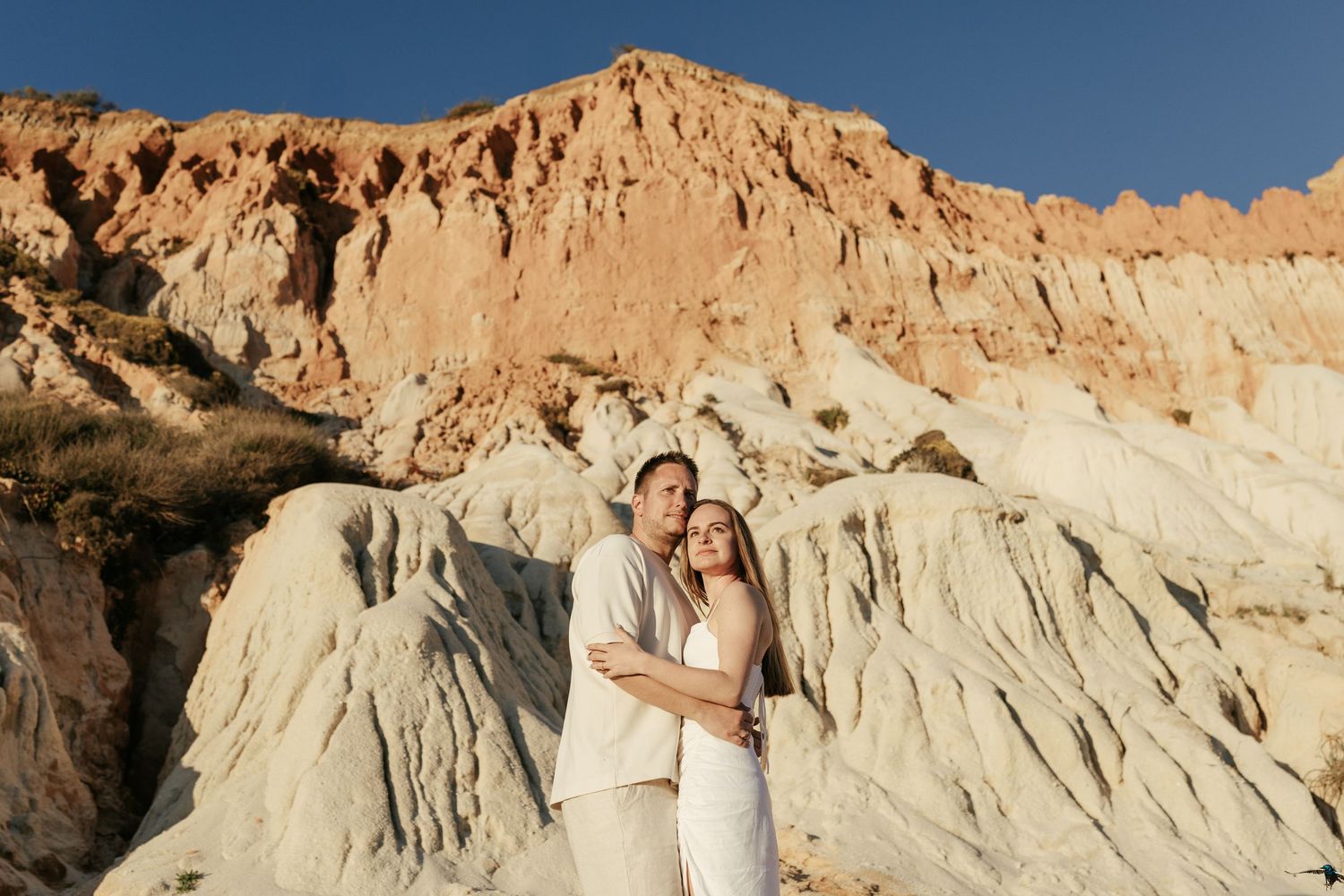 Two people embrace while standing against striking white and orange rock formations in a desert landscape at sunset.