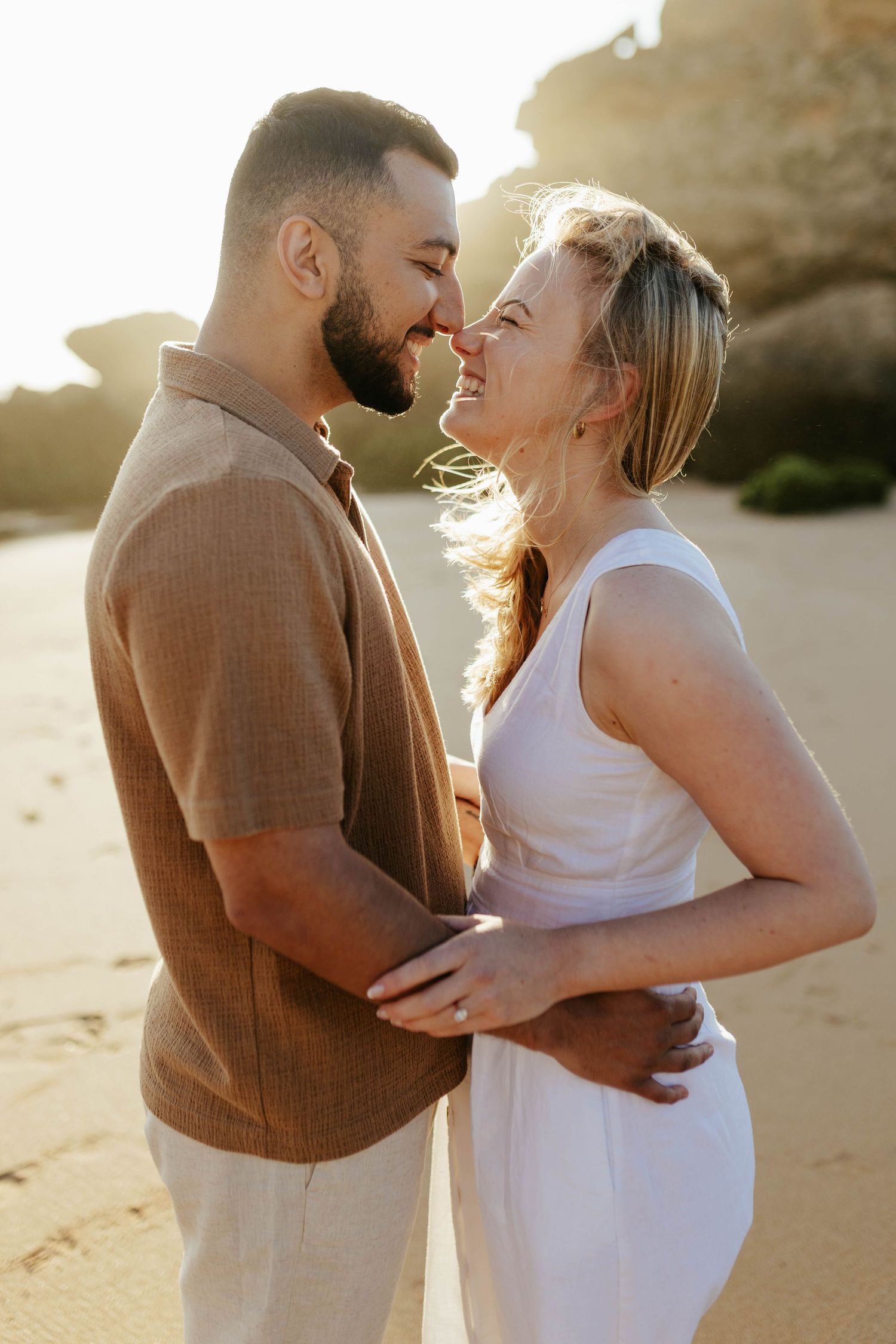 A romantic couple embraces on a sunlit beach at sunset, wearing light colored outfits against a golden backdrop.