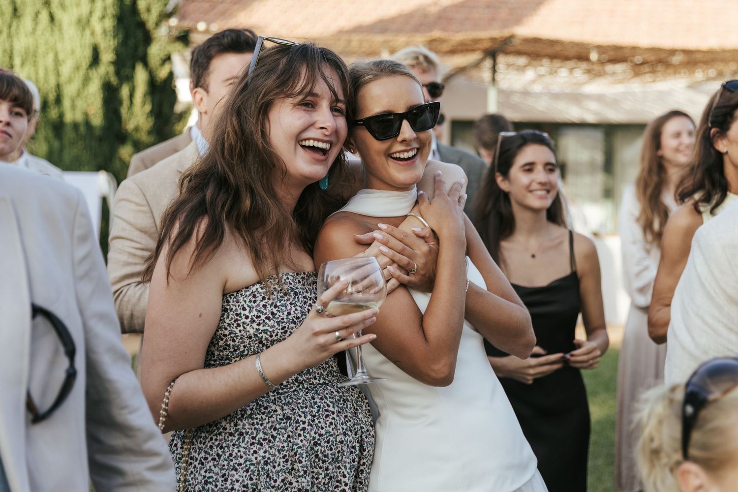 Bride in white dress laughing with female guest in floral dress at outdoor wedding.