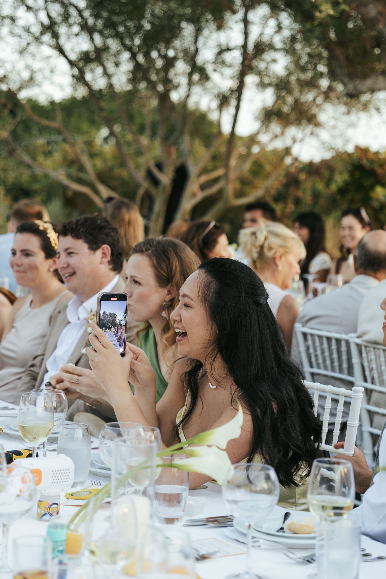 Guests laughing and taking photos on smartphone at elegant outdoor wedding reception dinner.