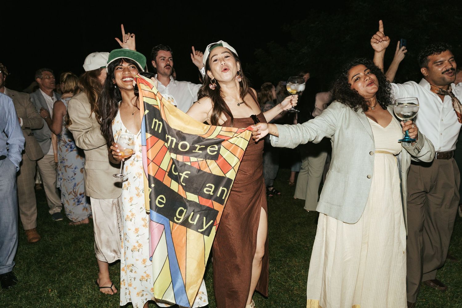 Three women dancing joyfully at outdoor wedding reception holding colorful banner at night.