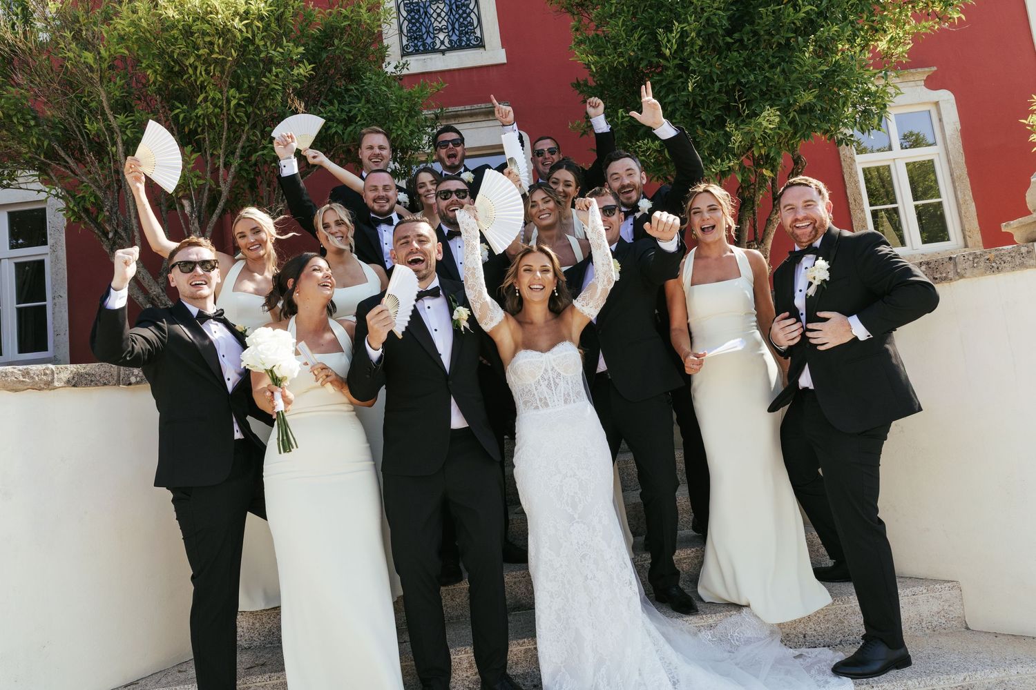 Wedding party celebrating outdoors in black tuxedos and white gowns, cheering joyfully near a red building.