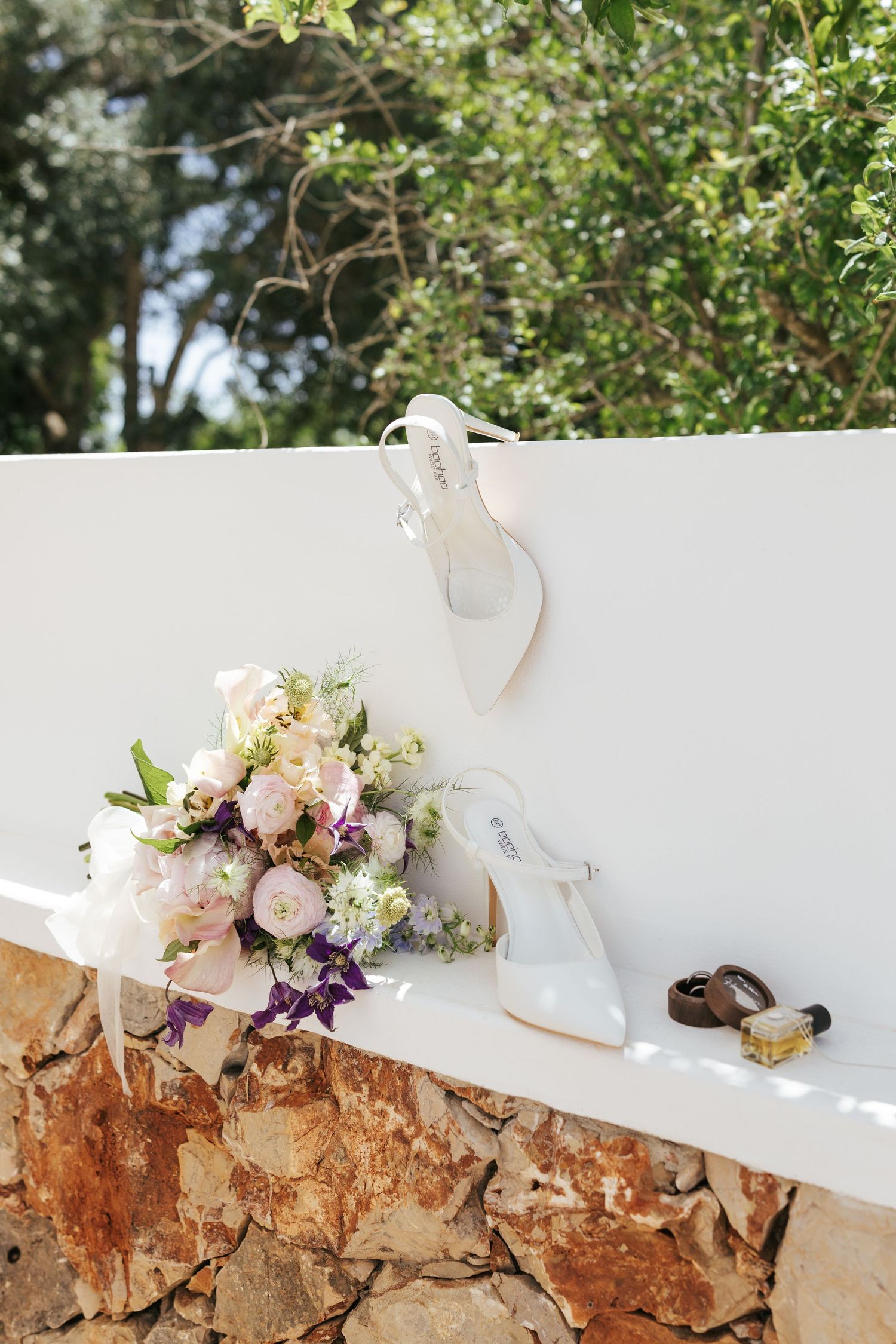 White bridal heels and floral bouquet with pink and purple flowers displayed on a stone wall outdoors.