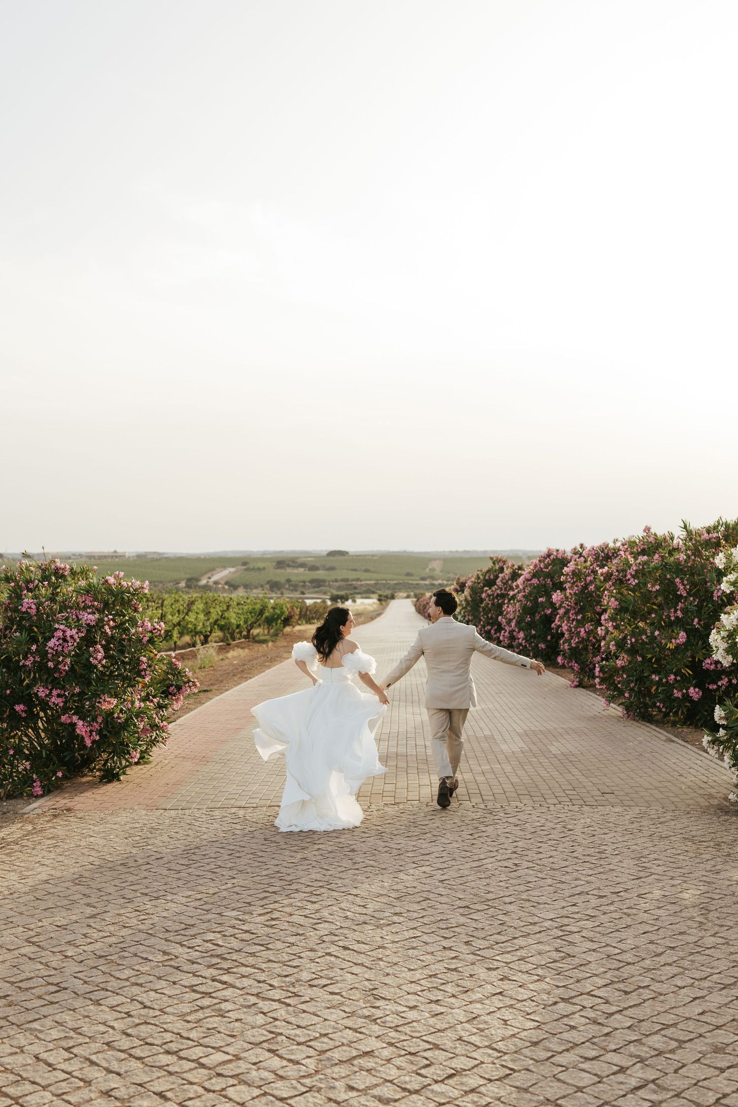 A couple in wedding attire playfully spins and dances on a stone path lined with blooming rose bushes.