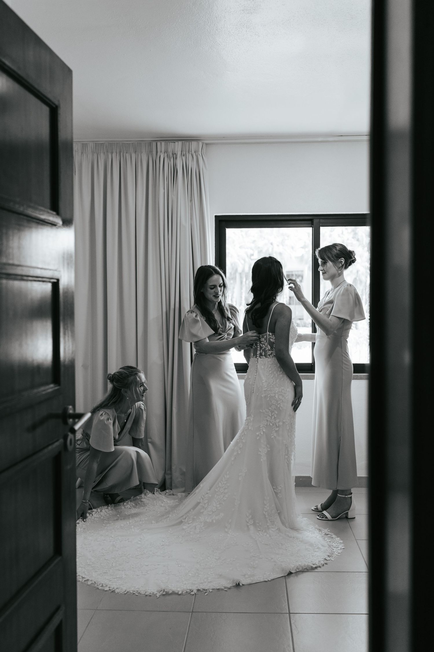 Black and white photo of a bride in a lace gown with bridesmaids helping her prepare in an elegant room.