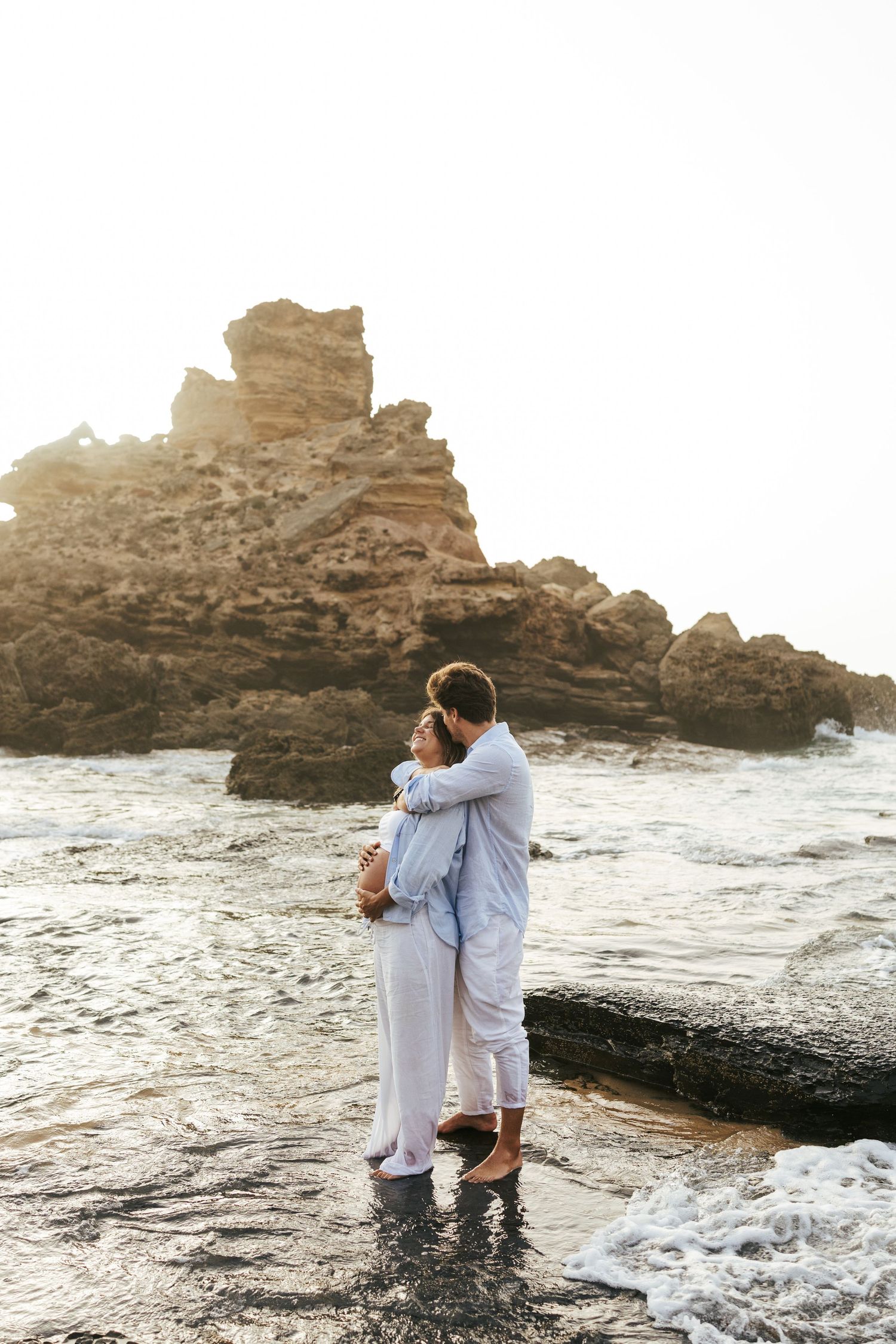 Couple embracing on a rocky beach with crashing waves and rugged cliffs in the background.