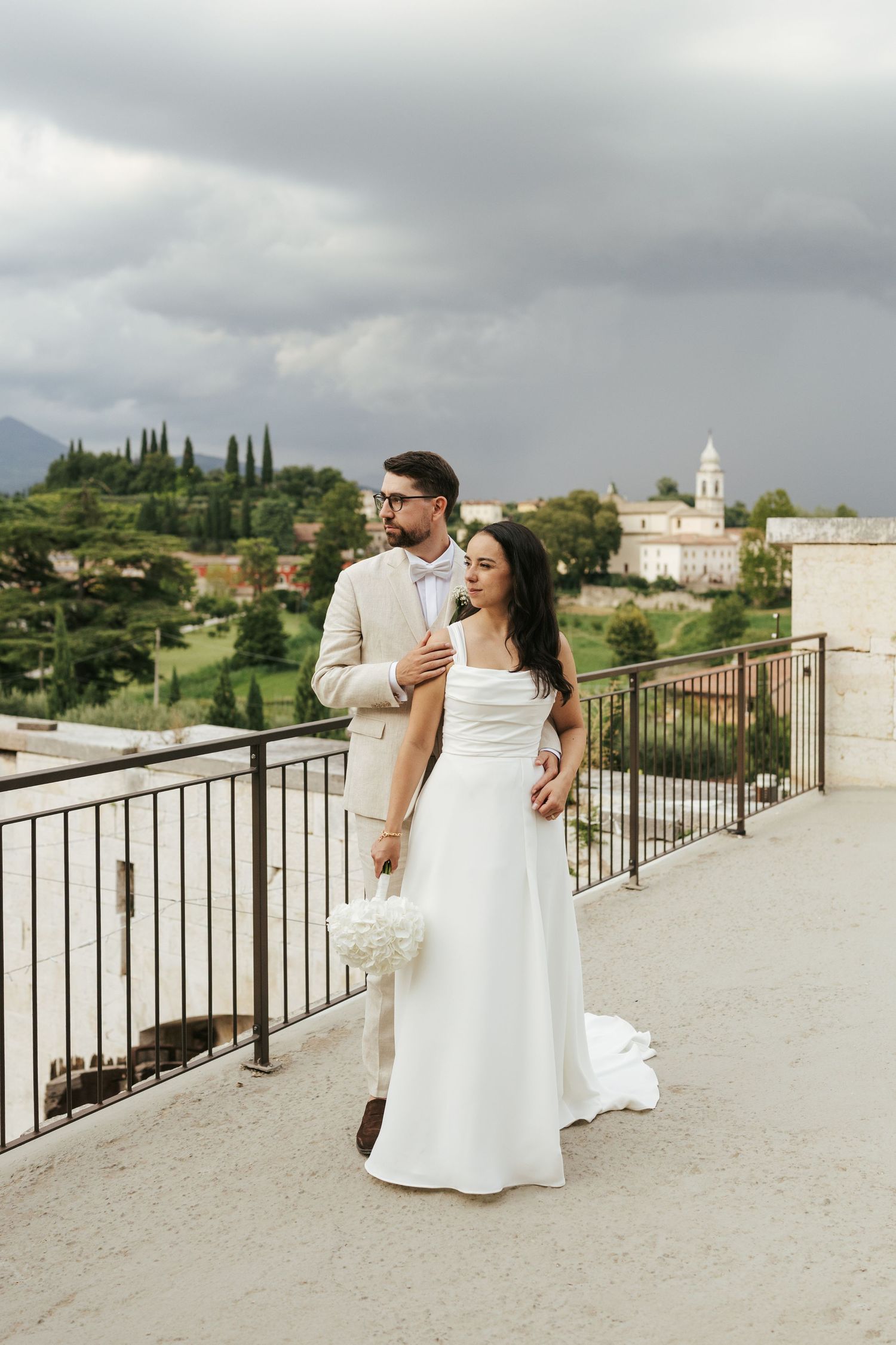 Wedding couple enjoying romantic moment on terrace overlooking Italian countryside and church.