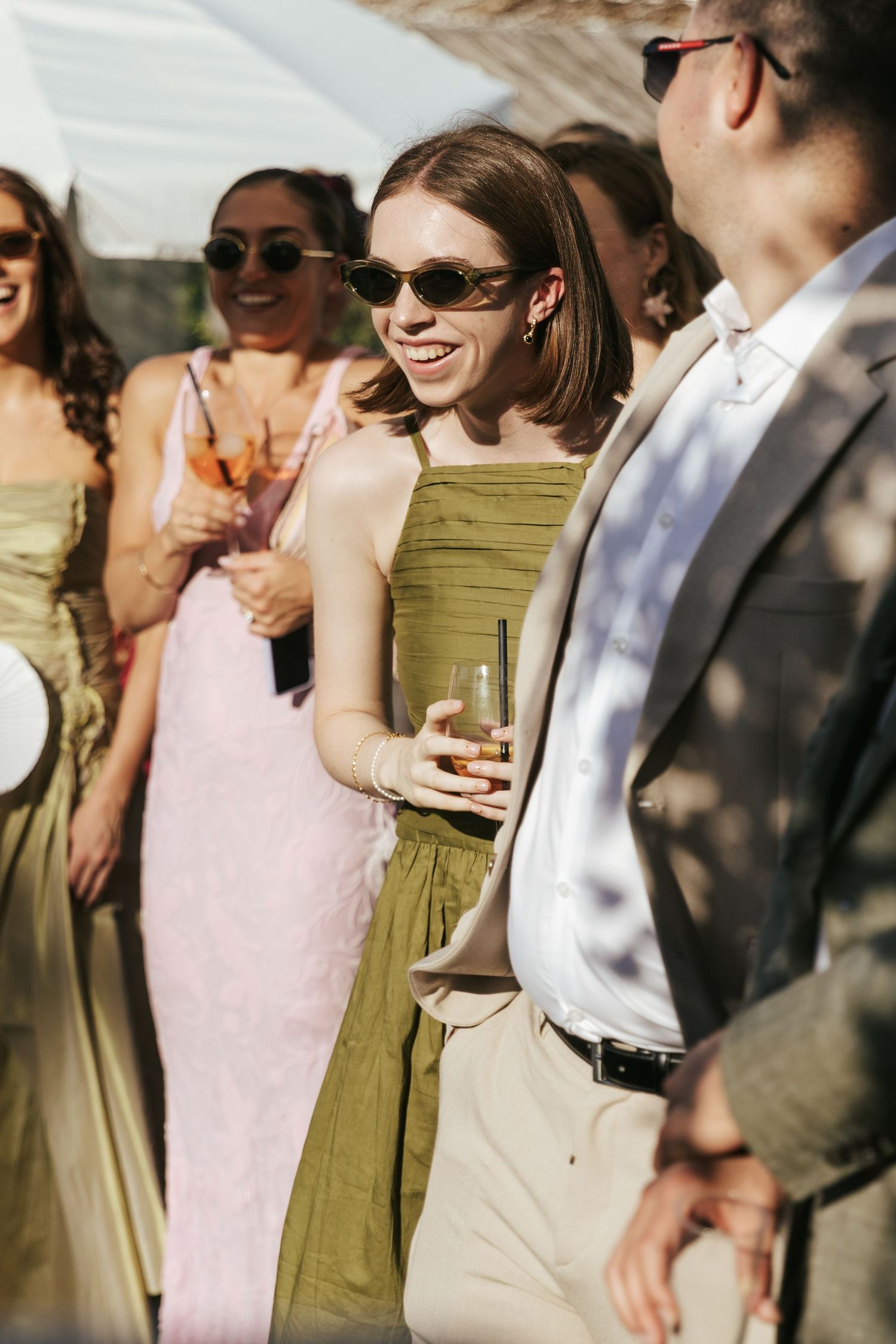 Woman in olive green dress and sunglasses smiling at outdoor wedding reception holding a glass.