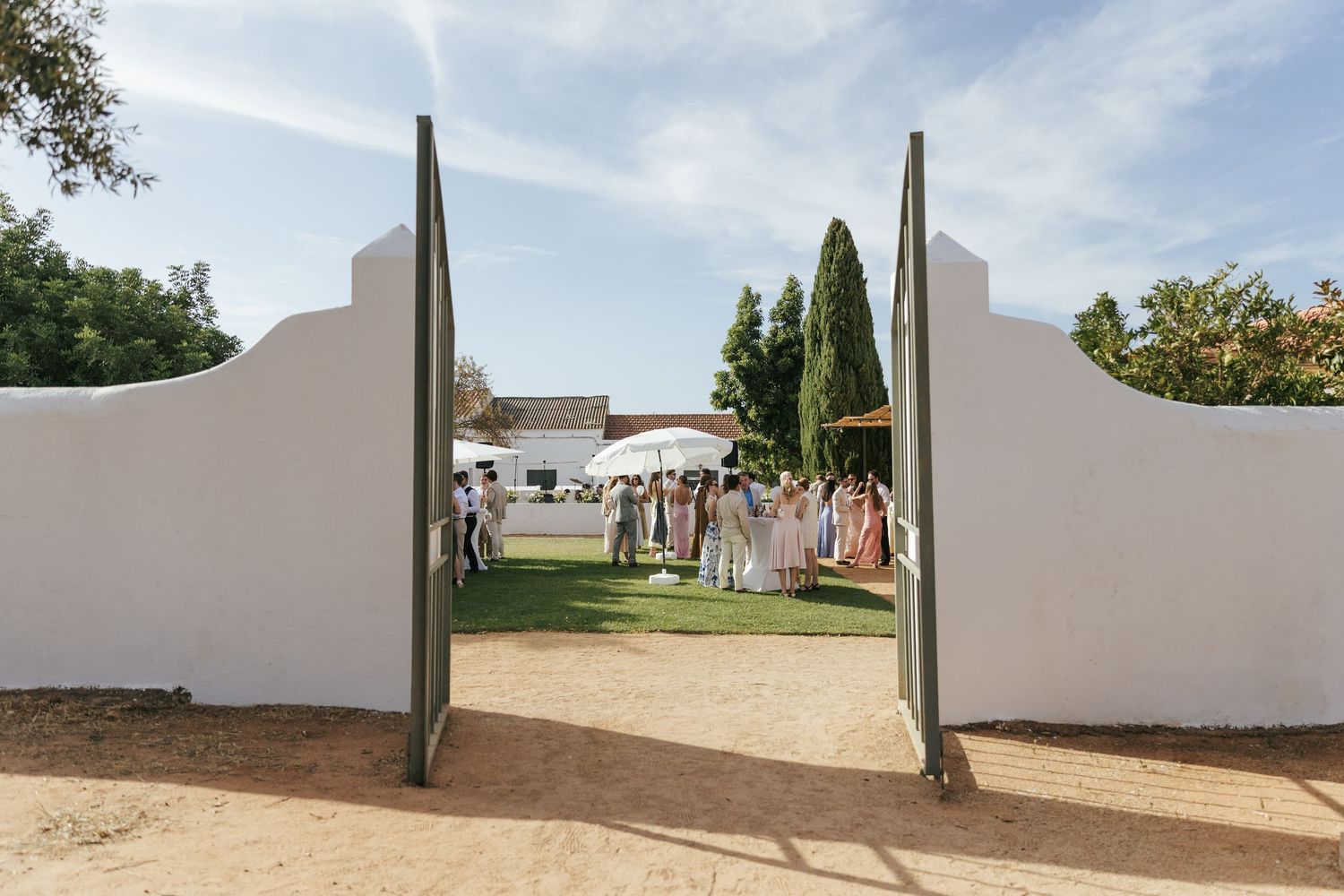 Elegant outdoor wedding venue with white walls and open gates revealing guests gathered on lush green lawn.