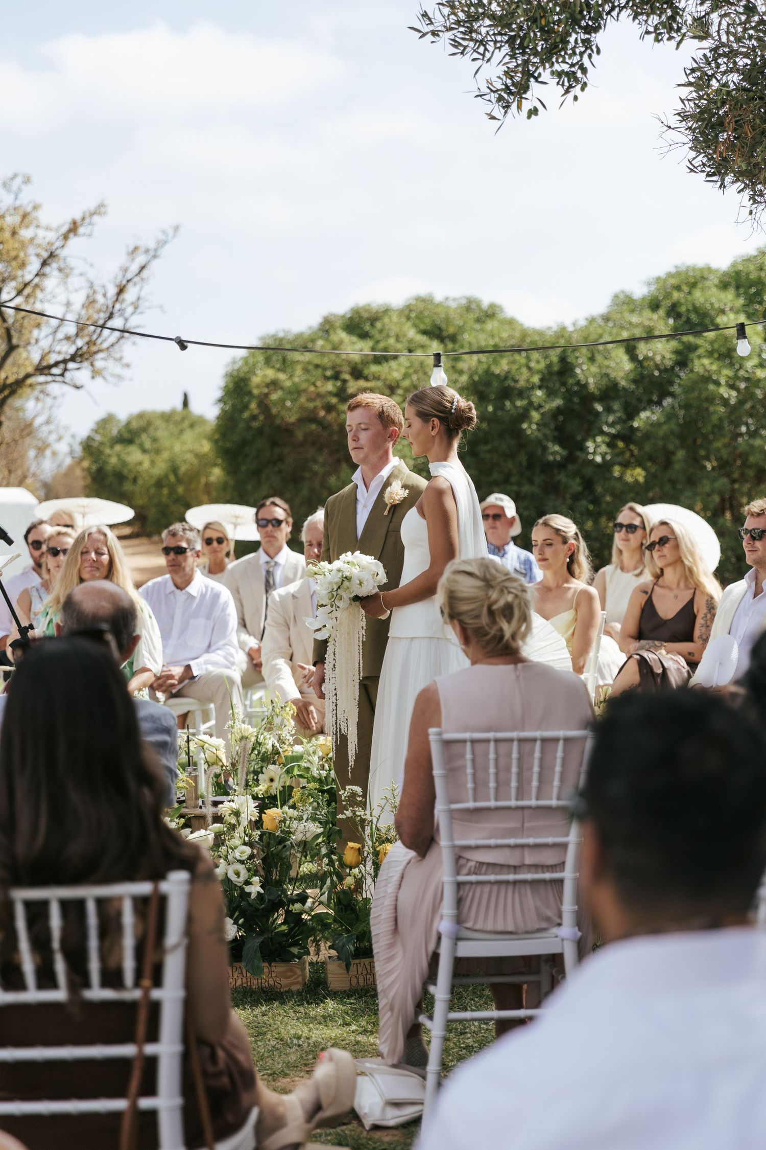 Bride and groom stand together during outdoor wedding ceremony surrounded by seated guests and floral arrangements.