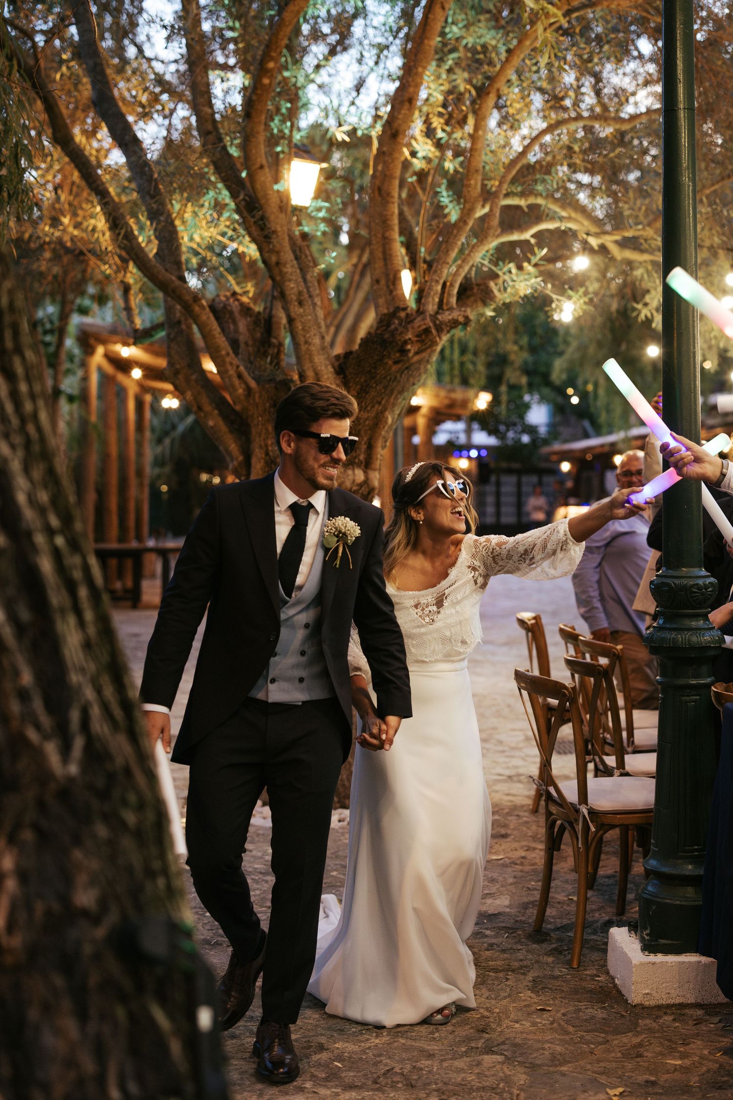 Newlywed couple wearing sunglasses dance together at an outdoor evening reception under string lights.