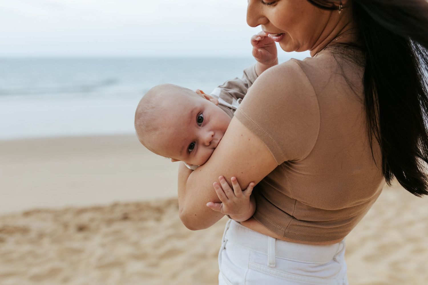 A parent cradles a small baby while standing on a sandy beach with the ocean in the background.