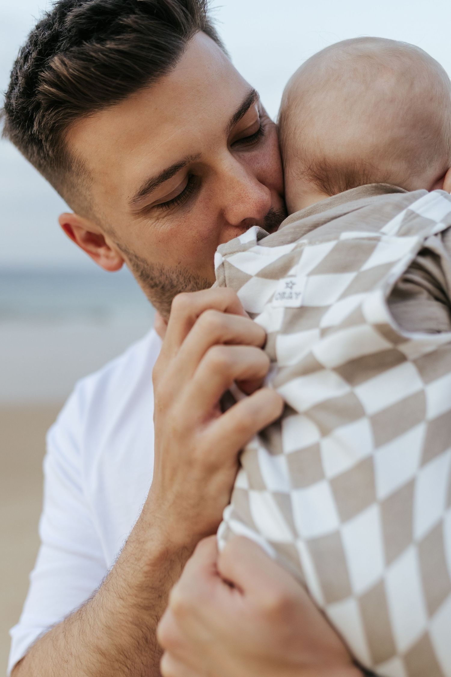 A parent in a white shirt tenderly kisses an infant wrapped in a checkered blanket at the beach.