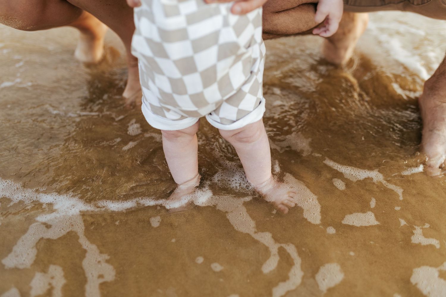 Small child's legs standing in shallow ocean water wearing checkered shorts as waves wash over their feet on sandy beach.