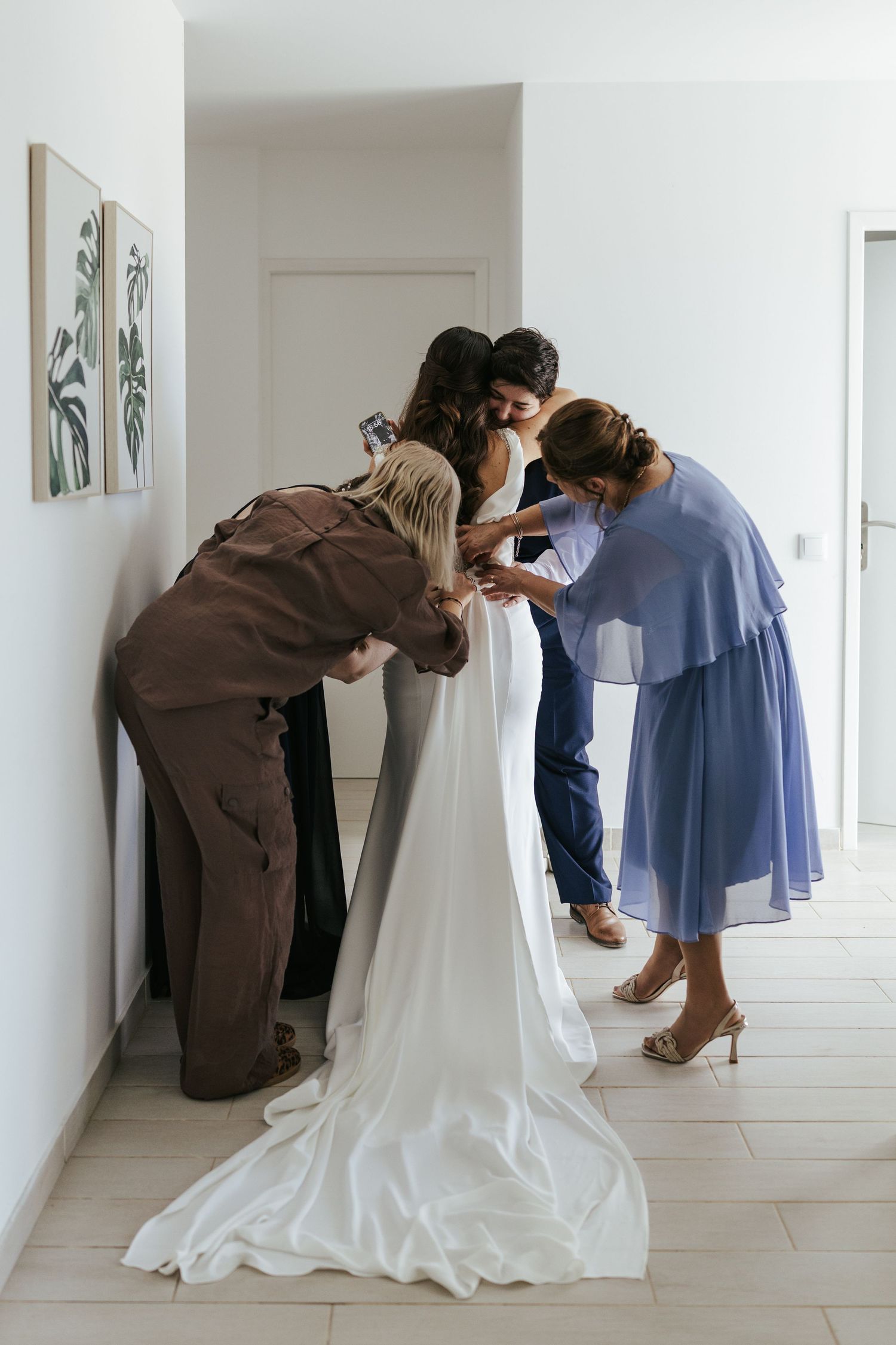 Helpers assist with adjusting a white wedding gown in a bright minimalist hallway with framed artwork.