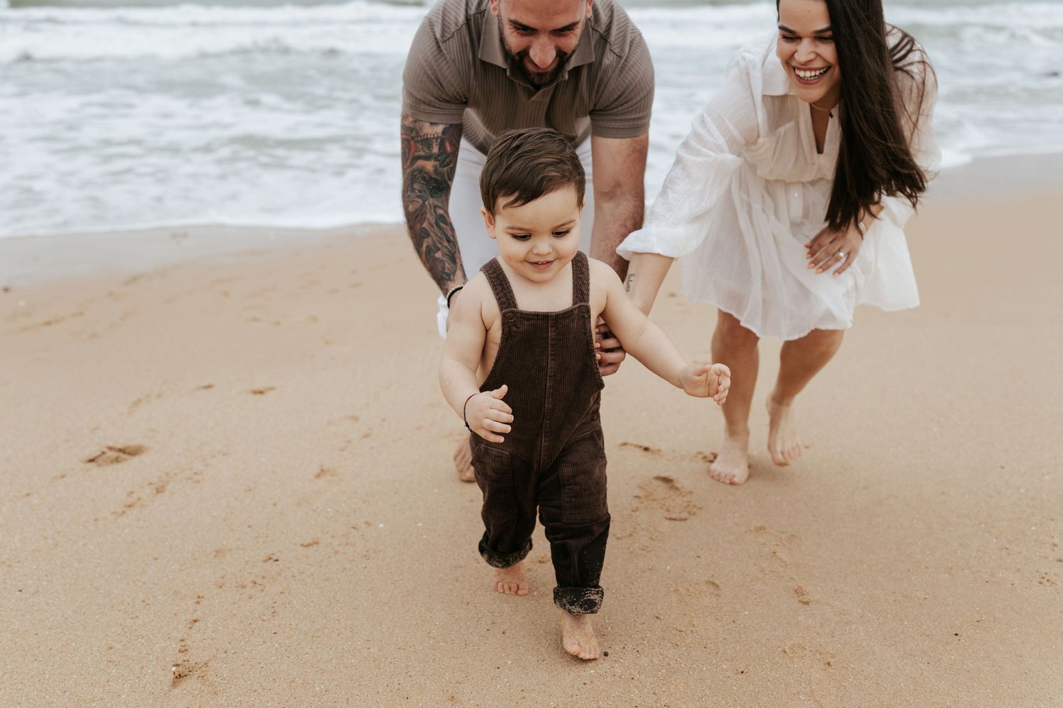 Family enjoys a walk together on sandy beach with toddler in overalls learning to walk near ocean waves.