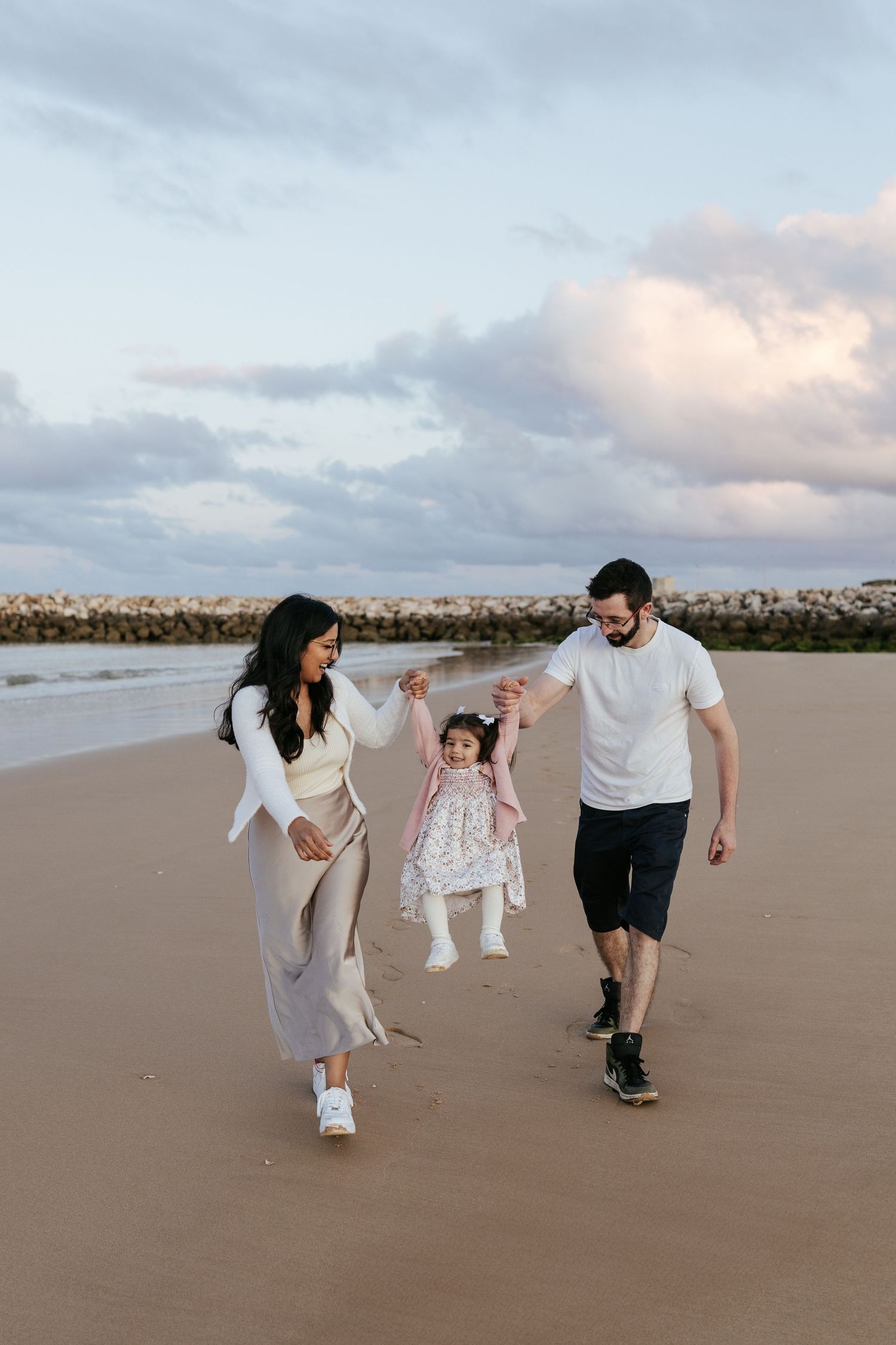 A family walks together on a sandy beach at sunset, swinging a small child between them while wearing casual attire.