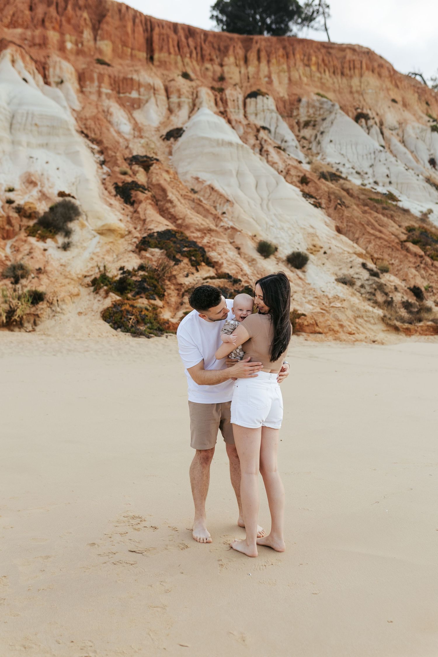 A couple embraces on a sandy beach with dramatic red and white striped cliffs in the background.