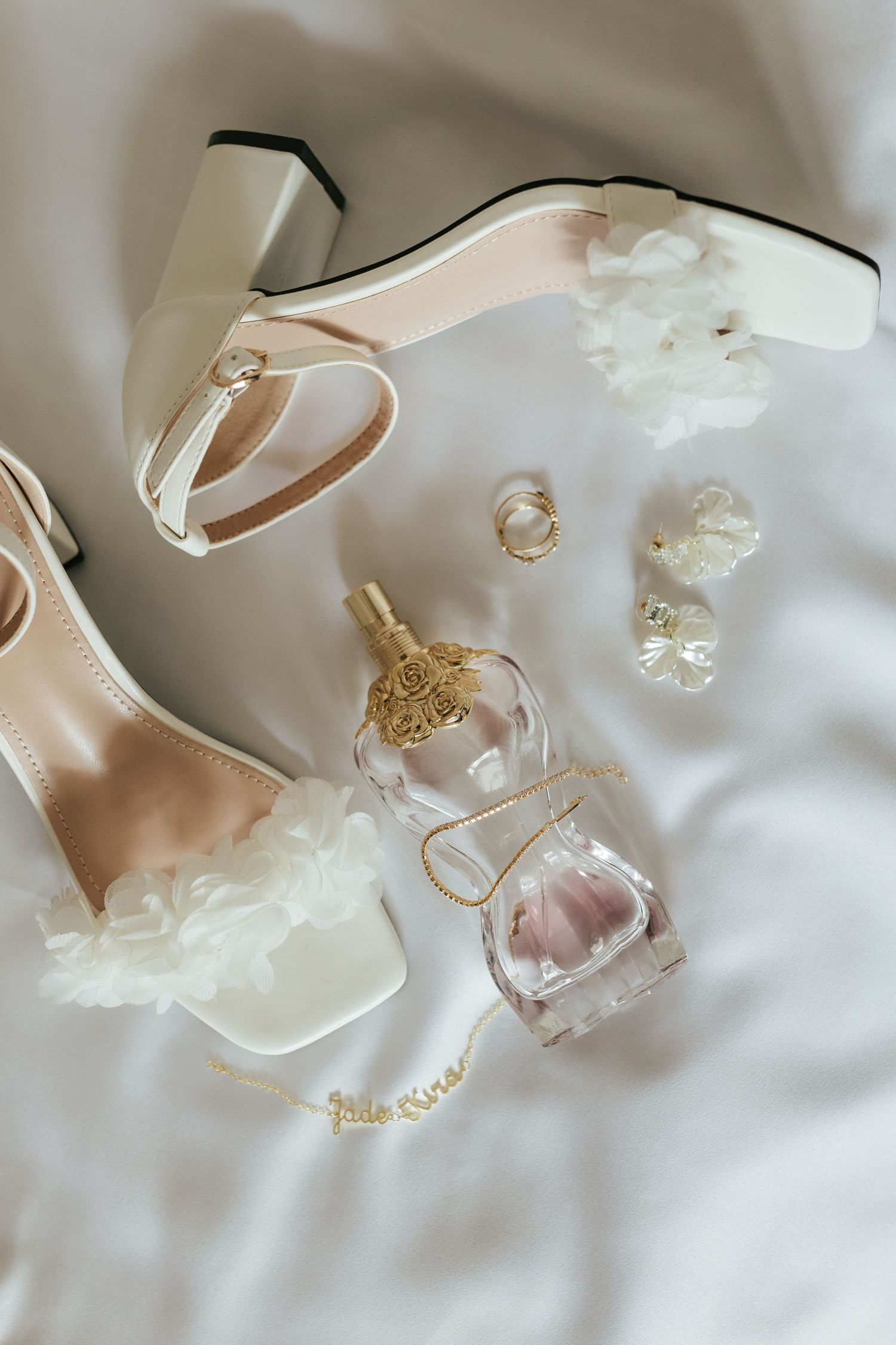 Wedding accessories including elegant white heels, perfume bottle, and jewelry arranged on white silk fabric background.