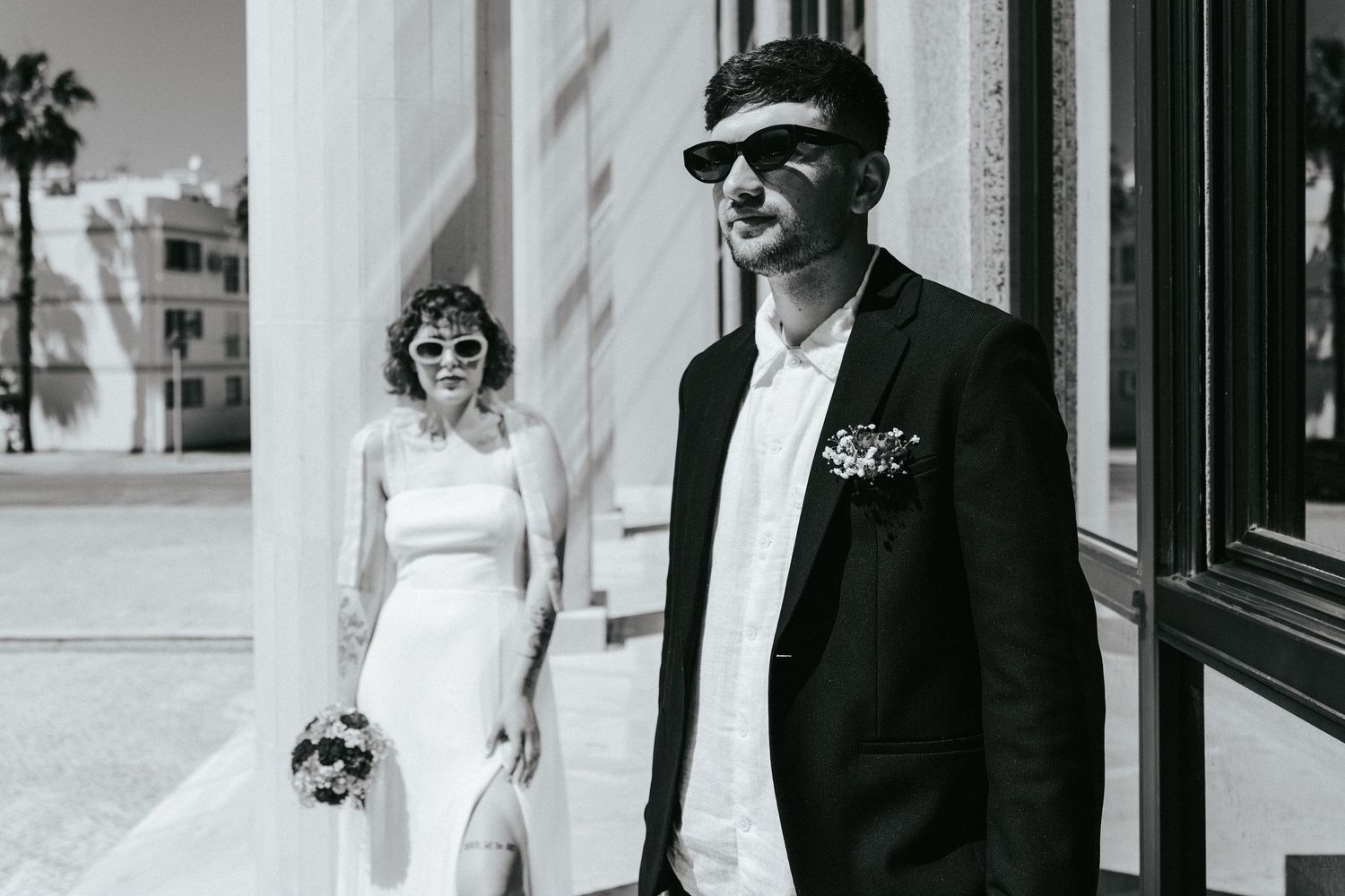 Black and white photo of a bride in white dress and groom in dark suit with sunglasses standing outside a building.