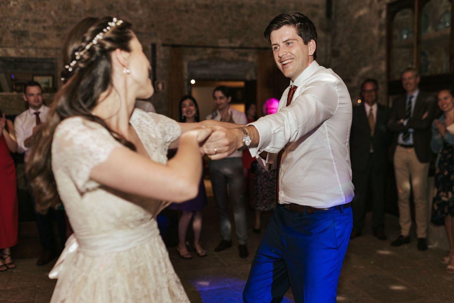 A couple dances together at their wedding reception, with the bride in a white dress and their guests watching in the background.