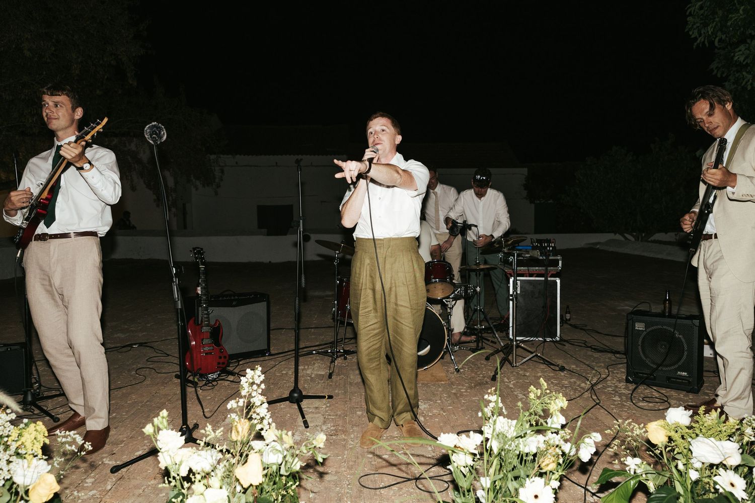 Outdoor band performing at night with musicians playing violin, singing, and guitar near floral arrangements.
