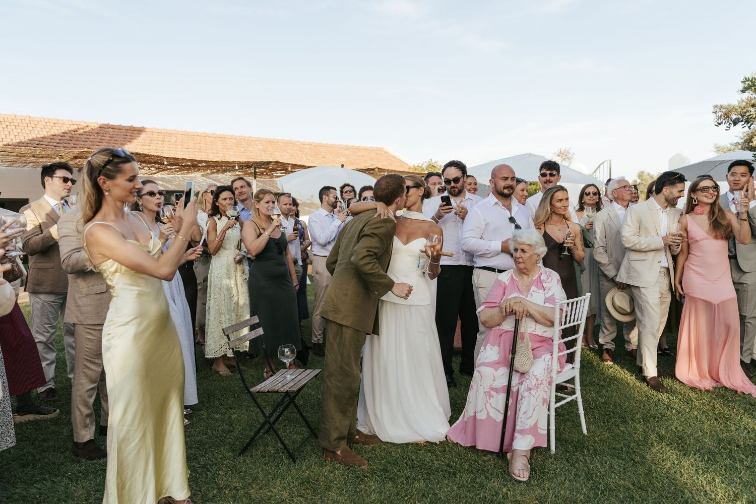 Wedding guests applaud and celebrate as a bride and groom share a moment together on a sunny outdoor lawn venue.