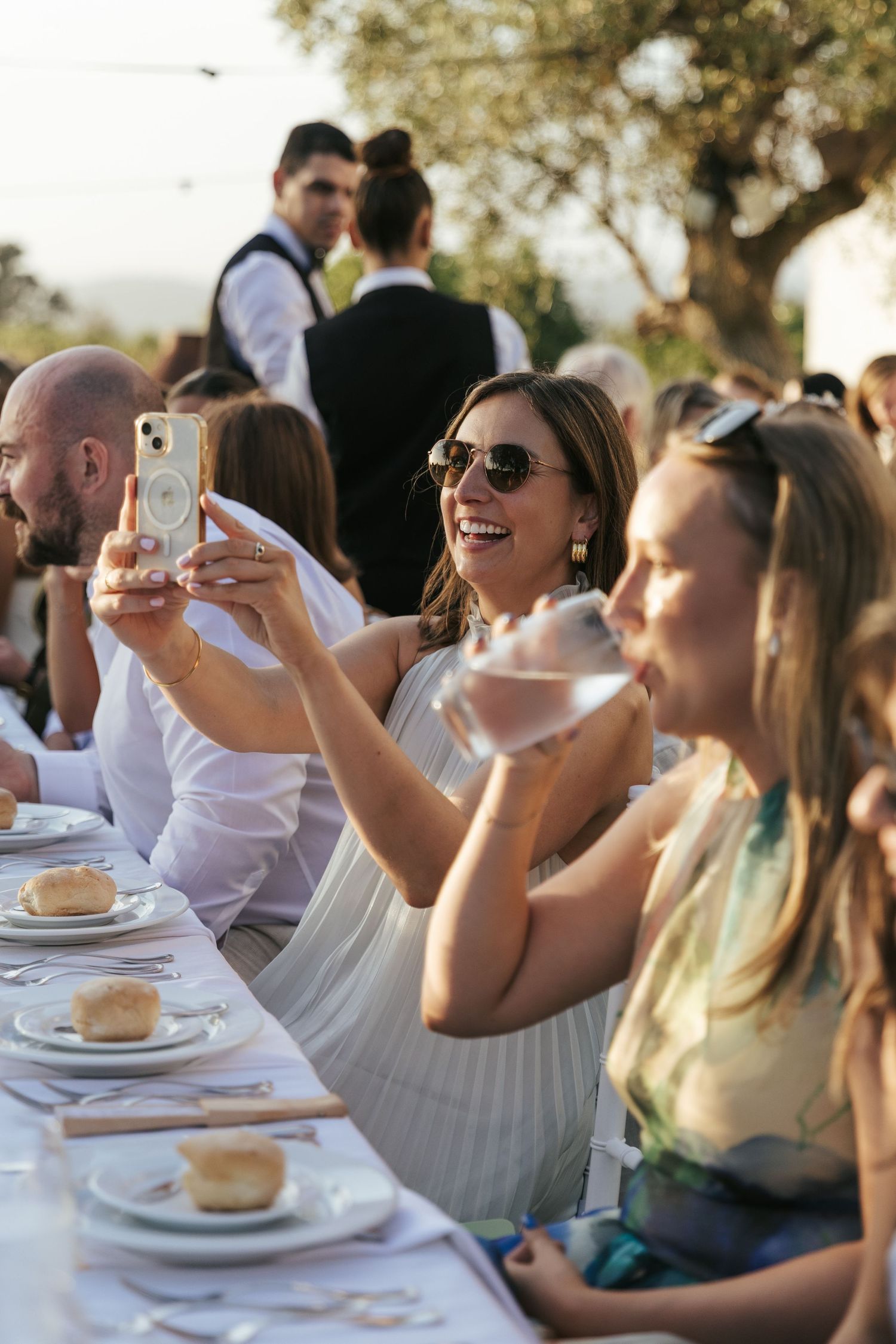 Smiling woman in sunglasses takes a photo on her phone at an outdoor wedding reception dinner.