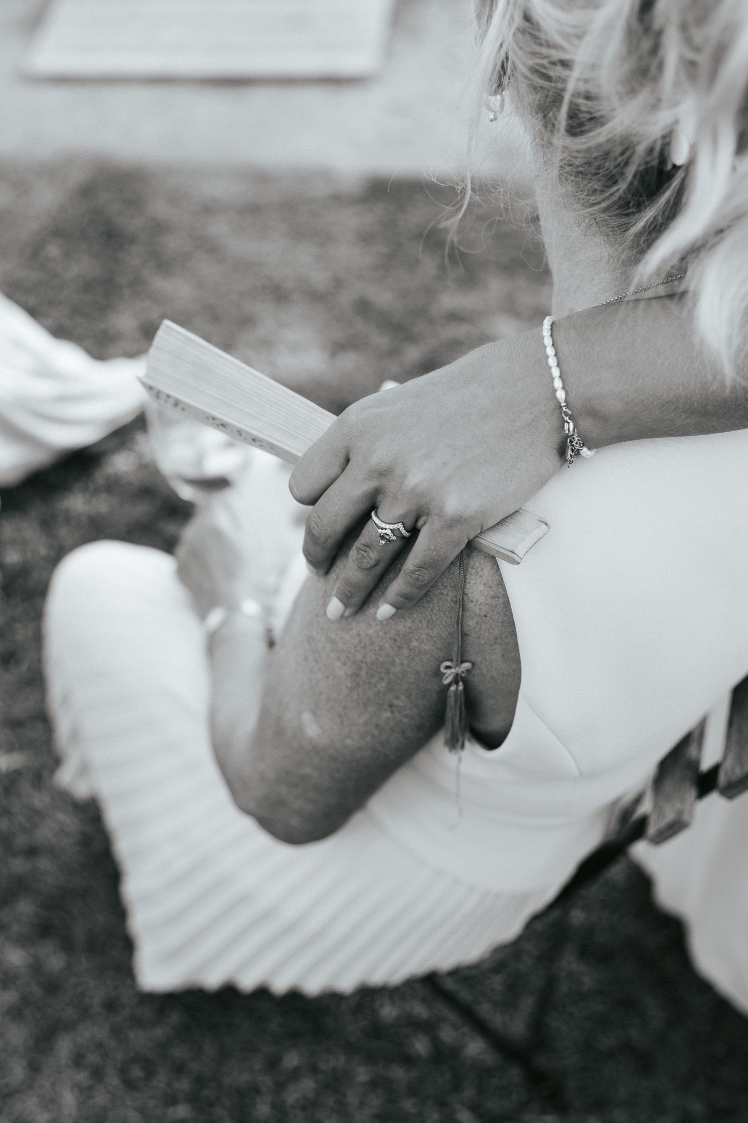 Black and white close-up of a woman in white dress holding a book on her lap outdoors.