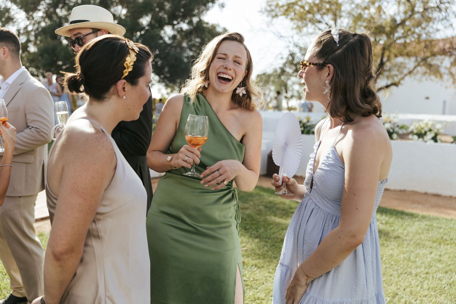 Three women laughing together at an outdoor wedding, one in a green dress holding a cocktail glass.