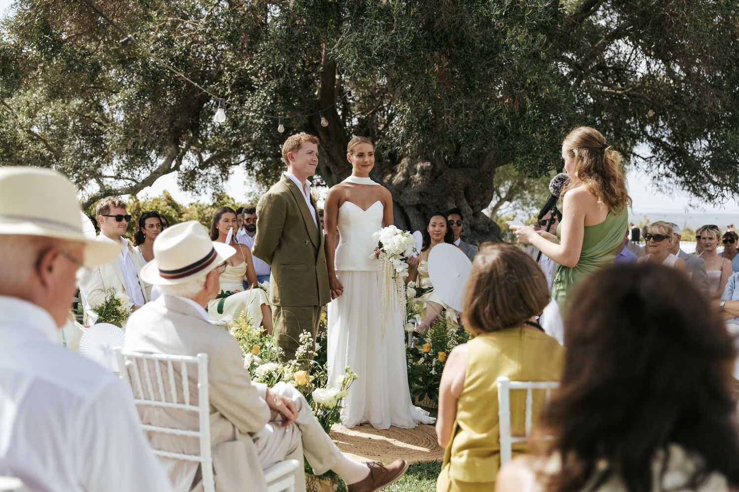 Outdoor wedding ceremony under olive tree with bride and groom standing at altar, guests seated on white chairs in sunlight.