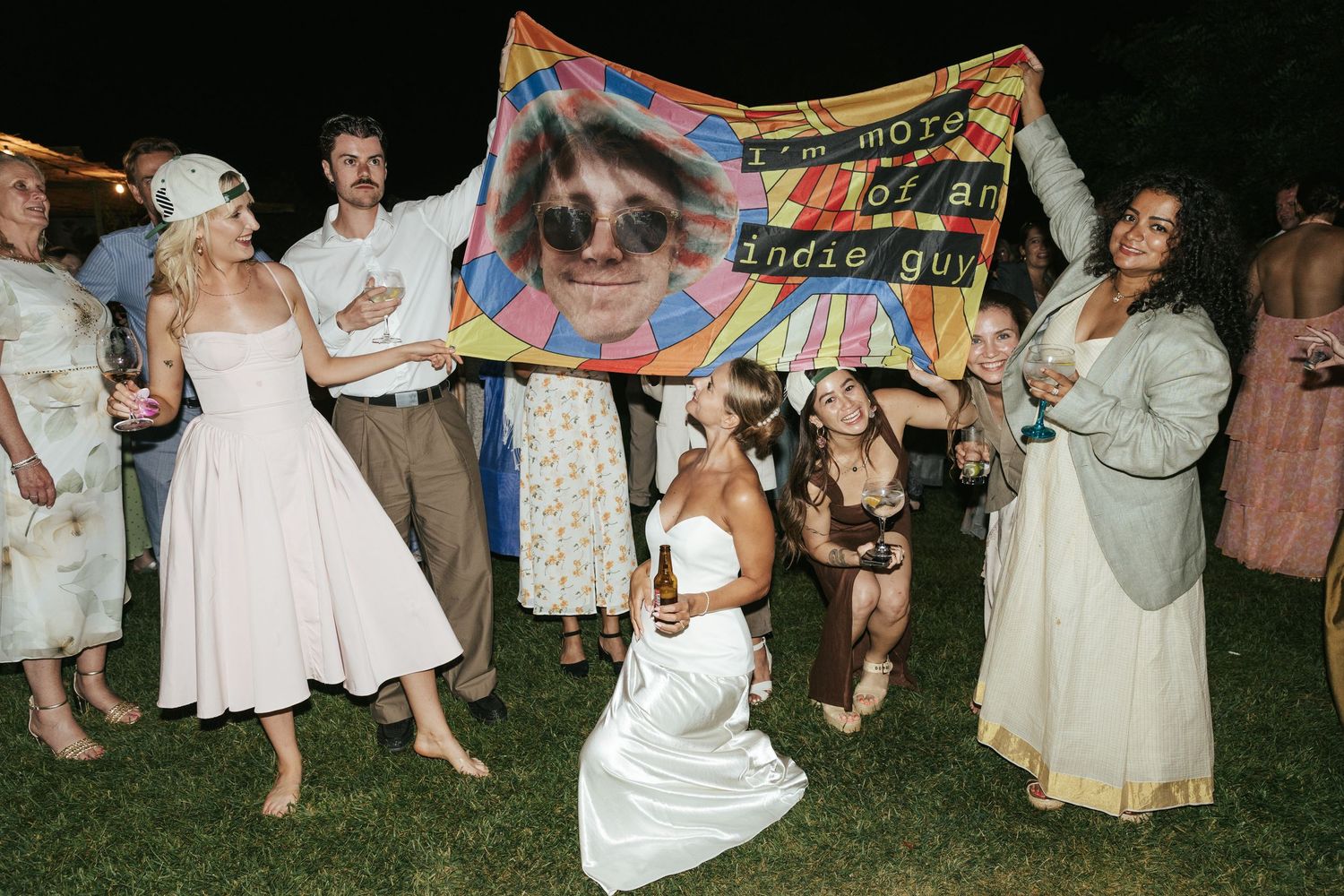 Wedding guests celebrate outdoors holding a colorful banner with a face print at a festive evening reception.