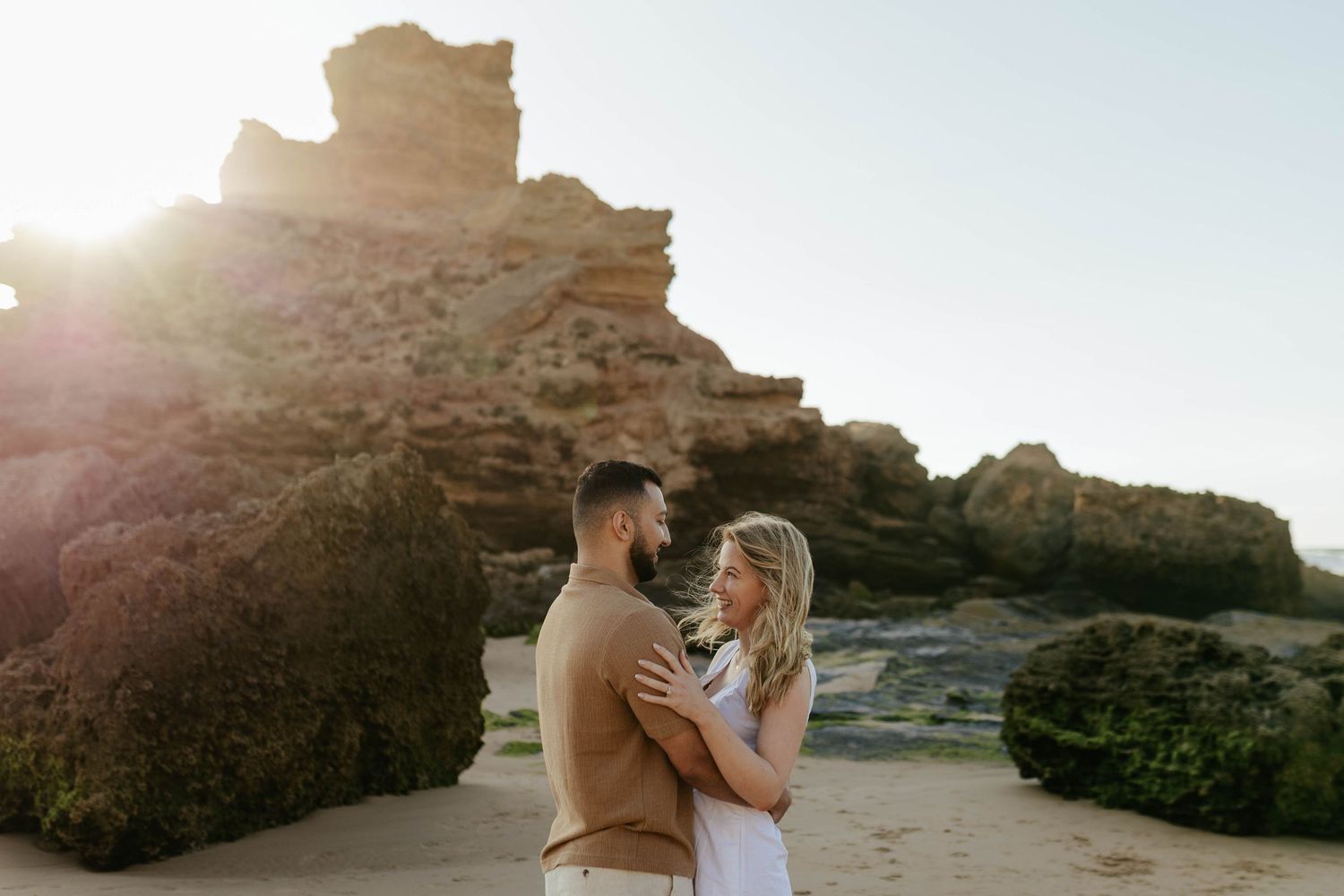 A romantic couple embraces on a scenic beach with towering rocky cliffs and golden sunlight.