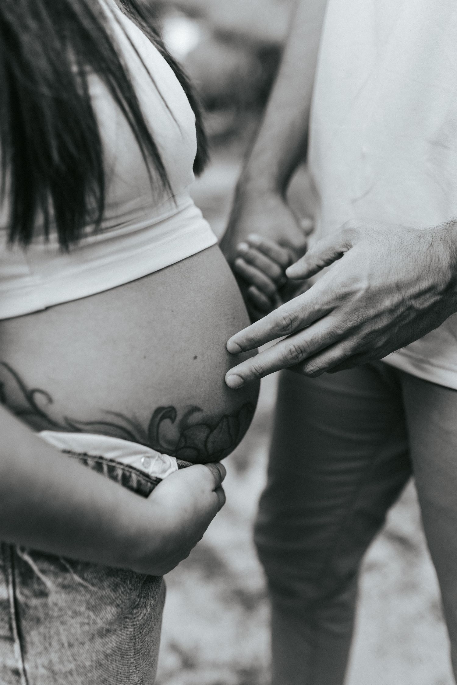 Black and white close-up photograph showing hands touching a tattooed pregnant belly in an intimate maternity moment.