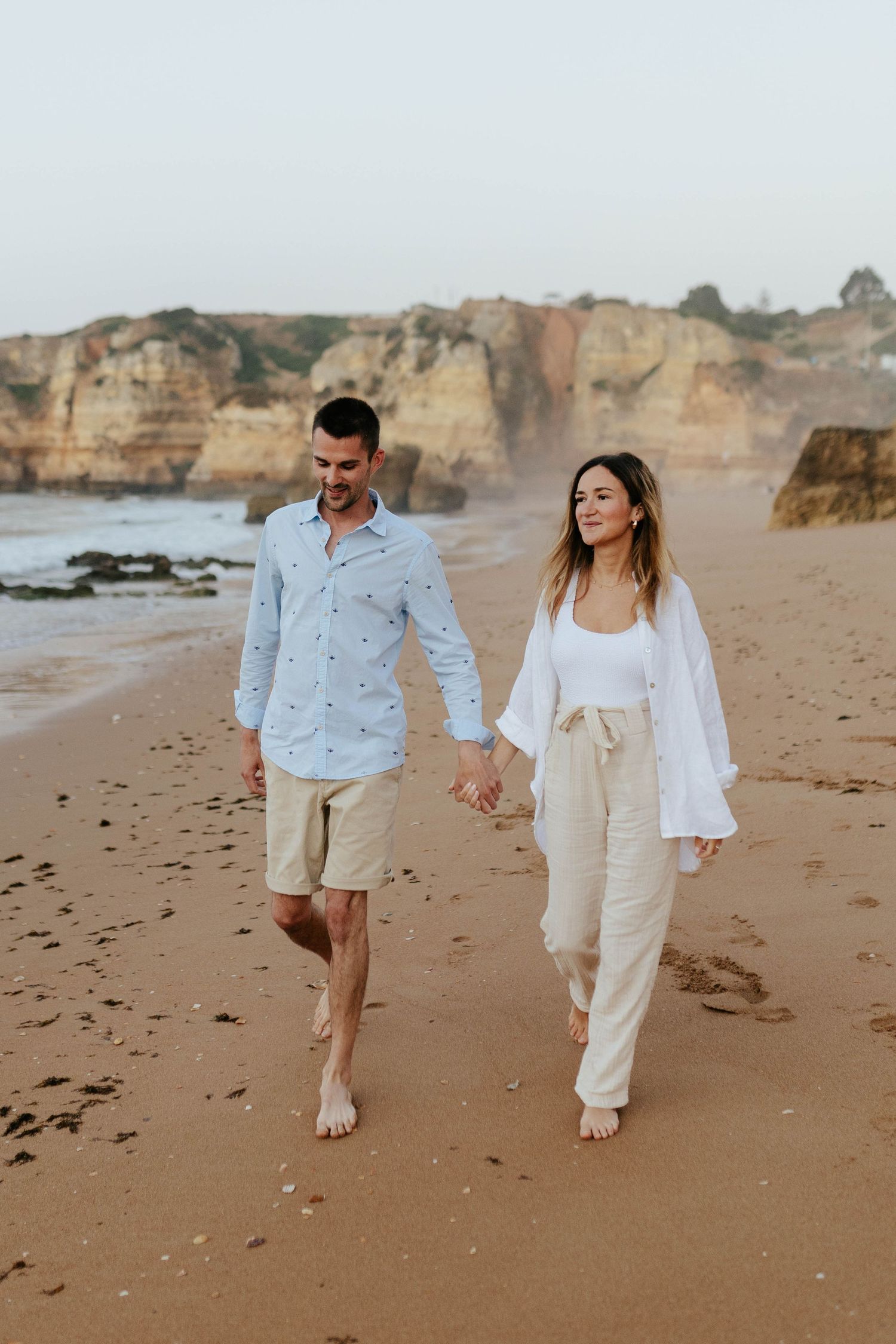 A couple takes a barefoot stroll along a sandy beach with dramatic coastal cliffs. Praia Dona Ana, Lagos, Algarve, Portugal.